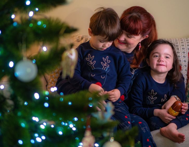 A family of three sit by a christmas tree. The mum and children are hugging each other and wear blue PJs