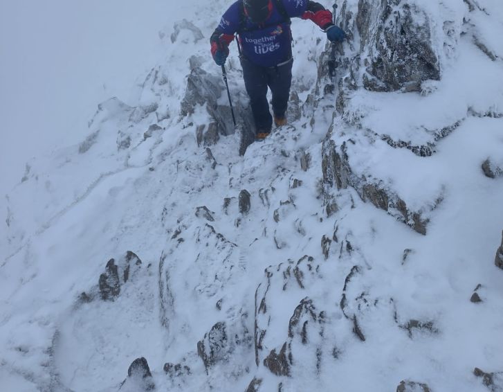 A man is trekking up a snowy, rocky mountainside. He has trekking poles in his hands and is wearing mountaineering kit as well as a Together for Short Lives purple sports-t-shirt.