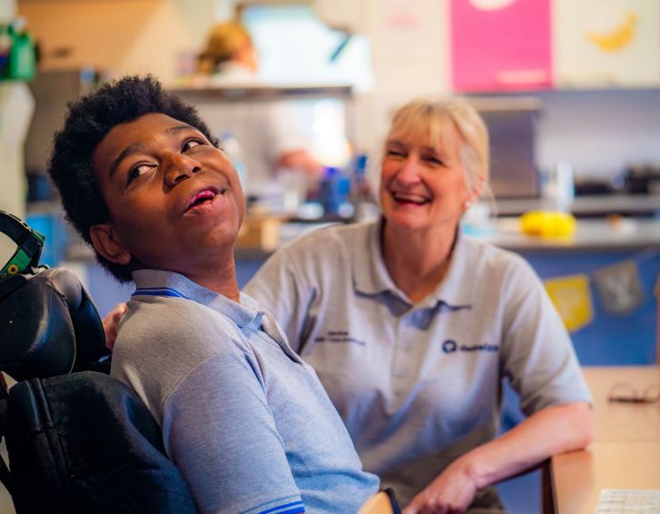 A young person sat in a wheelchair is smiling and looking at something behind the camera. A care professional sitting with him is also smiling.