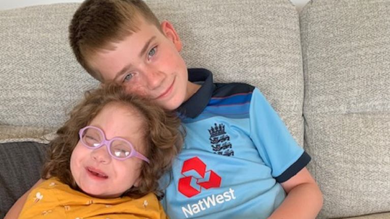 A young girl in a yellow jumper is pictured on a sofa with her brother, a young boy in a blue shirt.