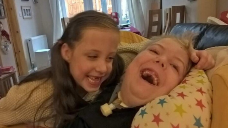A brother and sister are laughing together. the brother has a tracheostomy and is lying with a supportive neck pillow behind him.