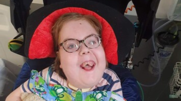 A young boy is seen lying down smiling up at the camera. He is wearing glasses.