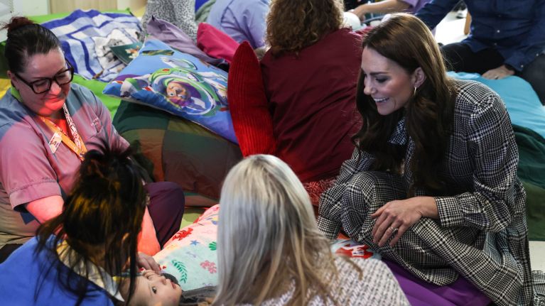 A room of people sitting down on the floor; in the foreground is Her Royal Highness The Princess of Wales smiling and interacting with a young girl. Surrounding the little girl are two children's hospice nurses and her mum.