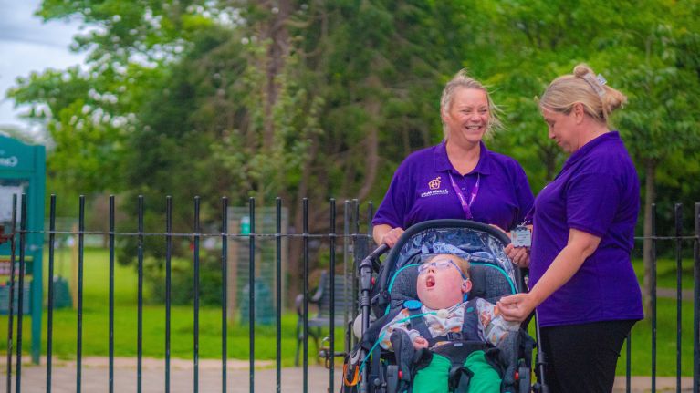 Two hospice nurses in purple t-shirts are stood by a park pushing a pram. In it is a young boy who is seriously ill.