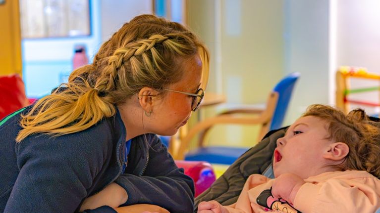 A palliative care nurses leans in to interact with a disabled girl laid back in a chair.