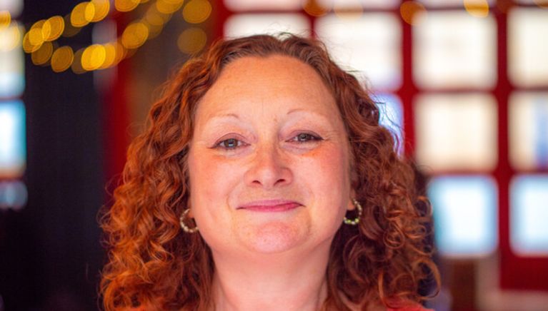 A white woman with curly red hair and a bright red top smiles directly at the camera