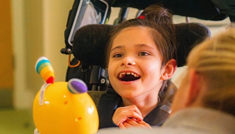 A child in a wheelchair smiles. They are looking at a brightly coloured toy, which is being show to them by a carer.