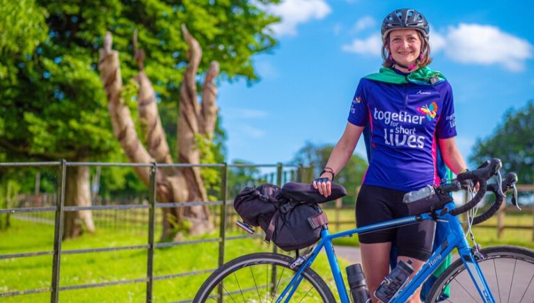 Francesca stands proudly with her blue bicycle. She's wearing a Together for Short Lives tshirt and a super hero cape.