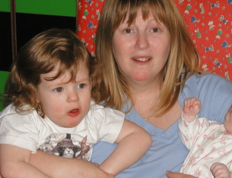 A mum is sat in a chair smiling at the camera. In each arm she cradles a child.