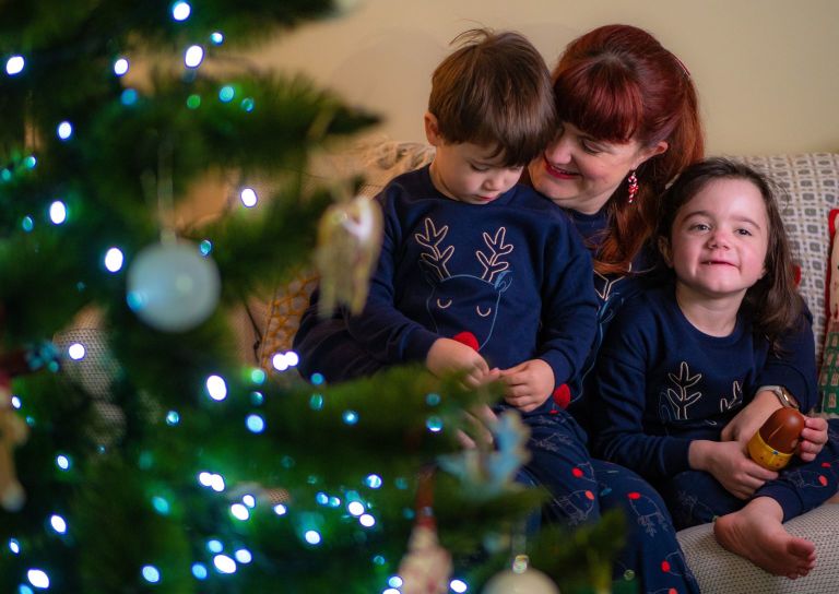 A family of three sit by a christmas tree. The mum and children are hugging each other and wear blue PJs