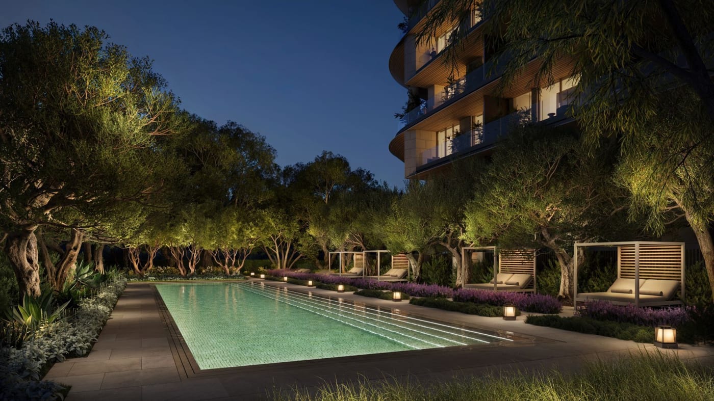 Rendu de la piscine des Aman Residences Beverly Hills la nuit, bordée d'oliviers et de cabanas, au pied d'une tour du complexe One Beverly Hills