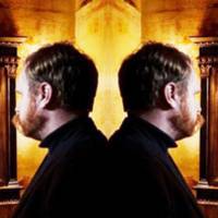 Image description: A mirrored image of Andy Slater standing back to back to himself, with his reflection duplicated into two mirrors on either side of the image. Tones of amber and yellow light emanate from the lights on either side of the mirrors. Slater is wearing a black turtleneck shirt.