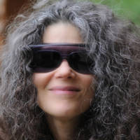 Image description: Claire is smiling. She has a long cascade of curly salt and pepper hair, and has on dark glasses and glare protecting fit-overs.