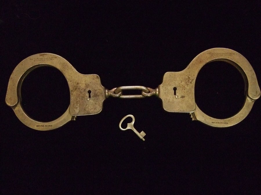 A photograph of a pair of silver handcuffs and a key against a black background