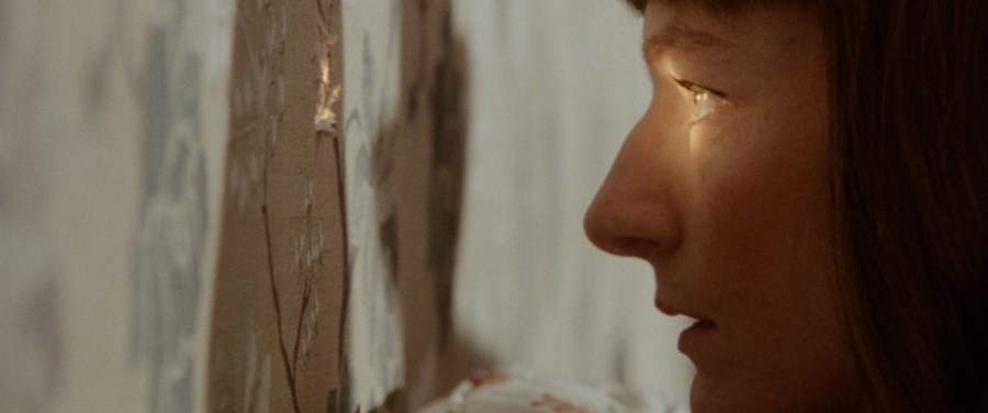 A woman peering through a small hole in the wall, through which a light shines on her face.