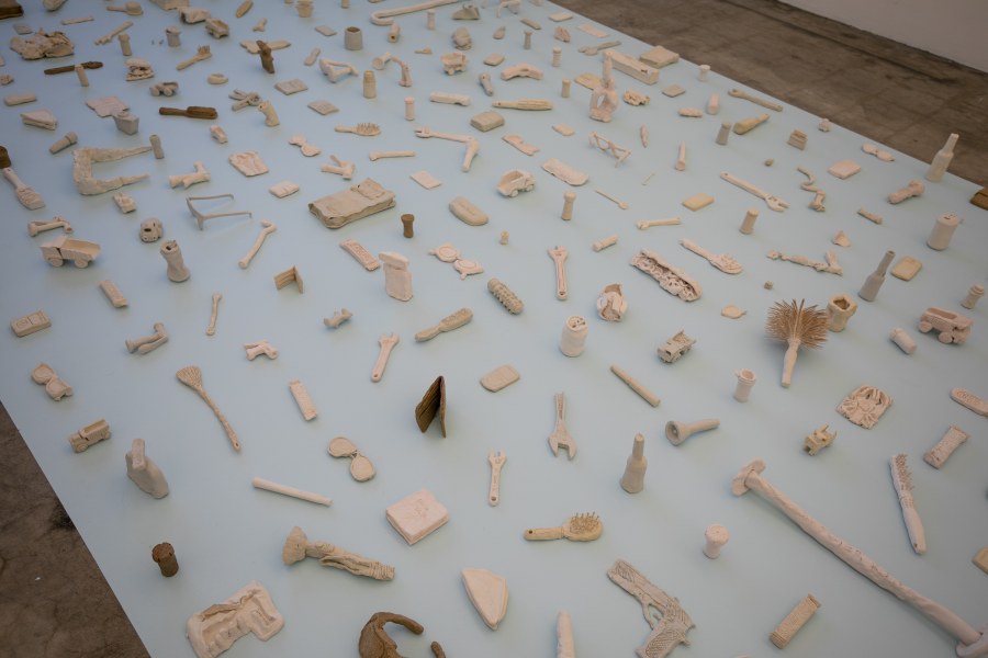 Numerous small, clay-colored sculptures of everyday objects sit on a pale blue platform.