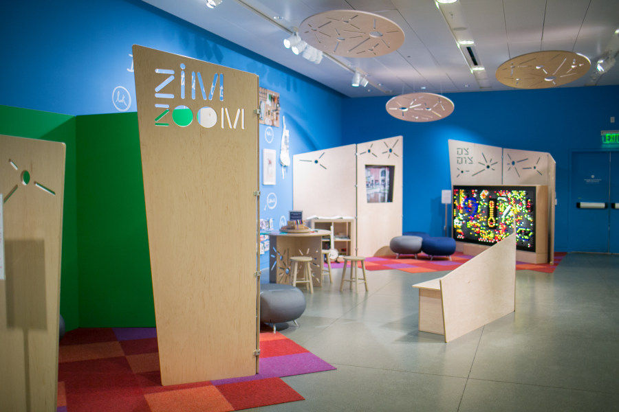 Installation view of the Zim Zoom Family Room. In the foreground, there is a large wooden panel with cut outs reading "Zim Zoom" with colorful carpets beneath it, and in the background there is an LED light wall with seating next to it..
