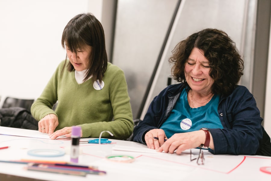 Two people sit at a table, smiling and working on paper crafts.