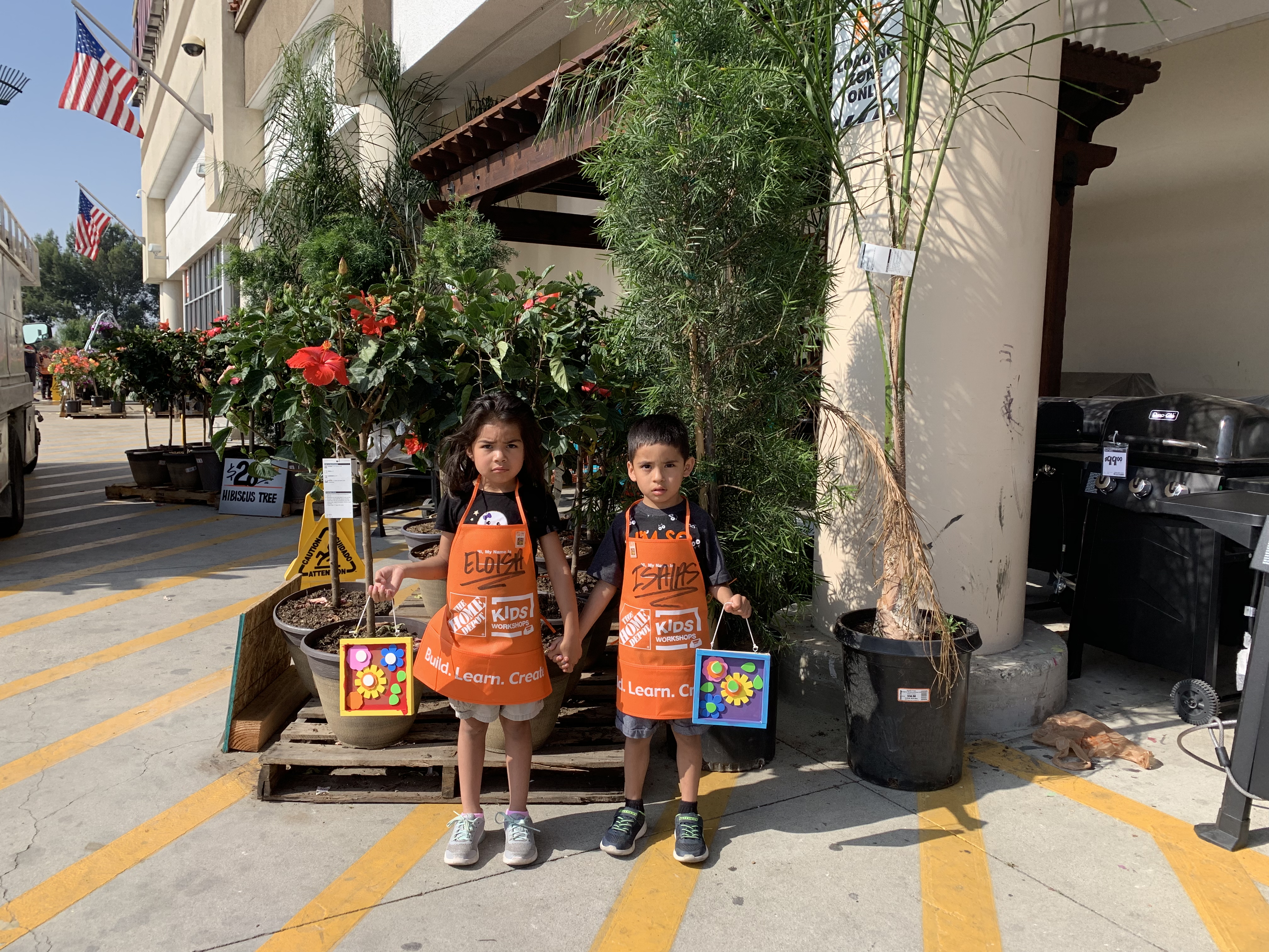 A color photo of two young children wearing orange Home Depot aprons, standing in front of a Home Depot