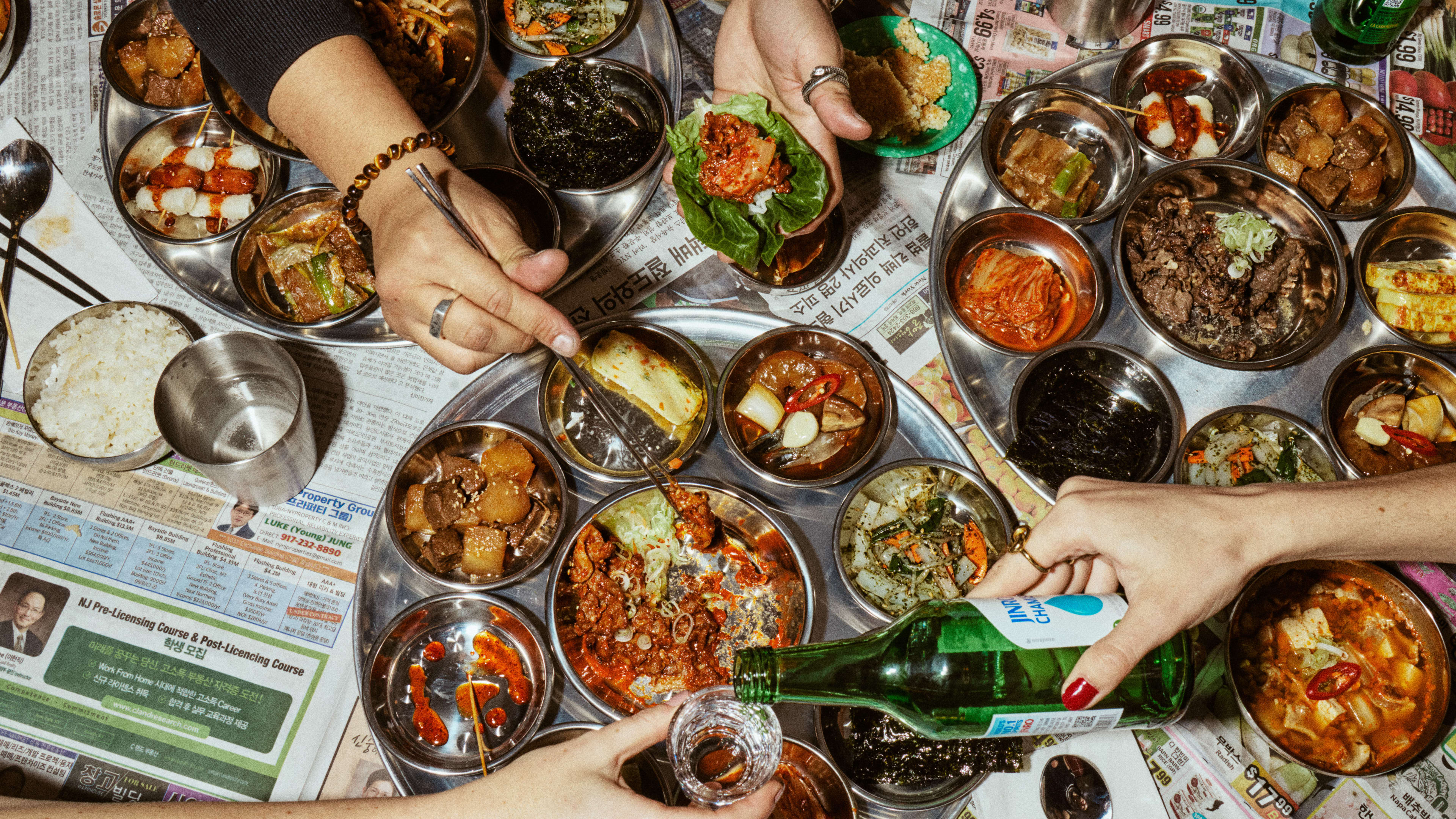 Flatlay of platters of bulgogi and spicy pork with banchan, hands with chopsticks and pouring soju.