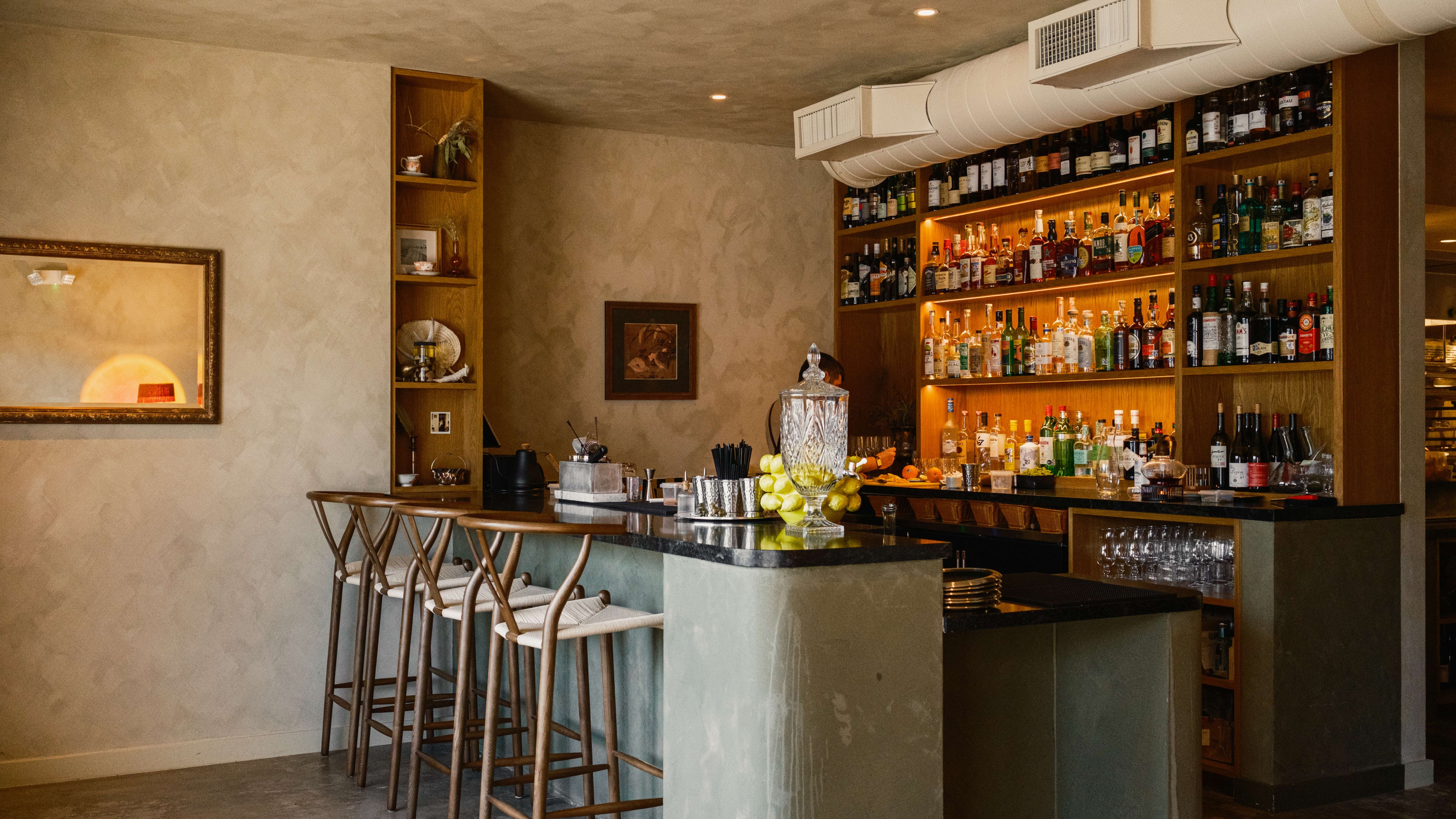 cocktail bar at Baso with a small bar top, four chairs, and an earthy/moody color palette with a back wall full of bottles