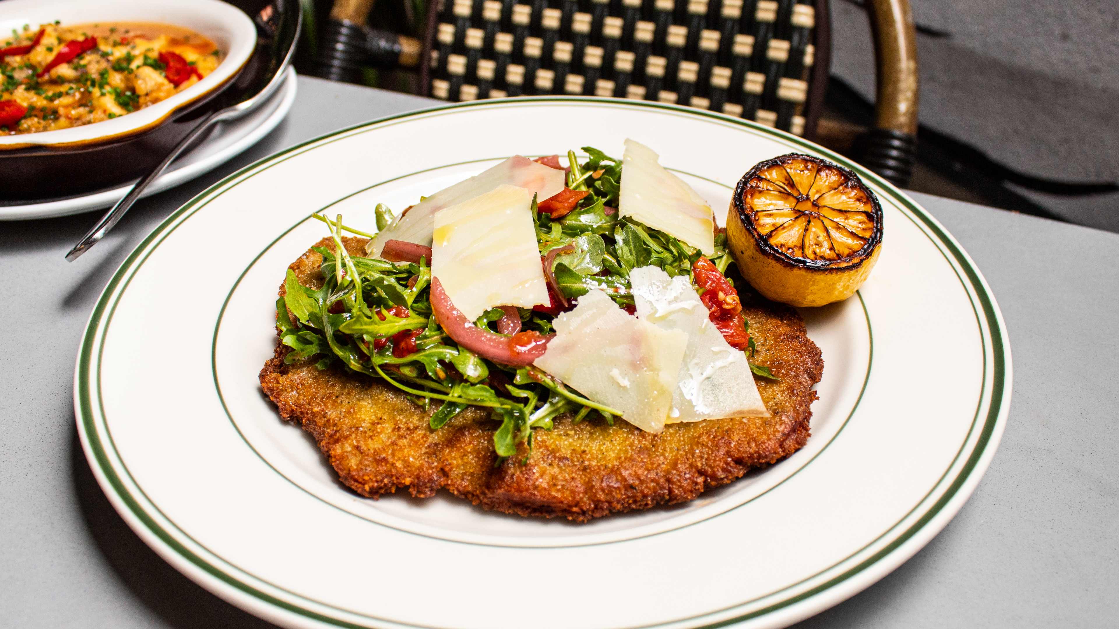 Breaded cutlet wtih arugula, tomatoes, shaved parm, and a burnt lemon half on a ceramic plate.