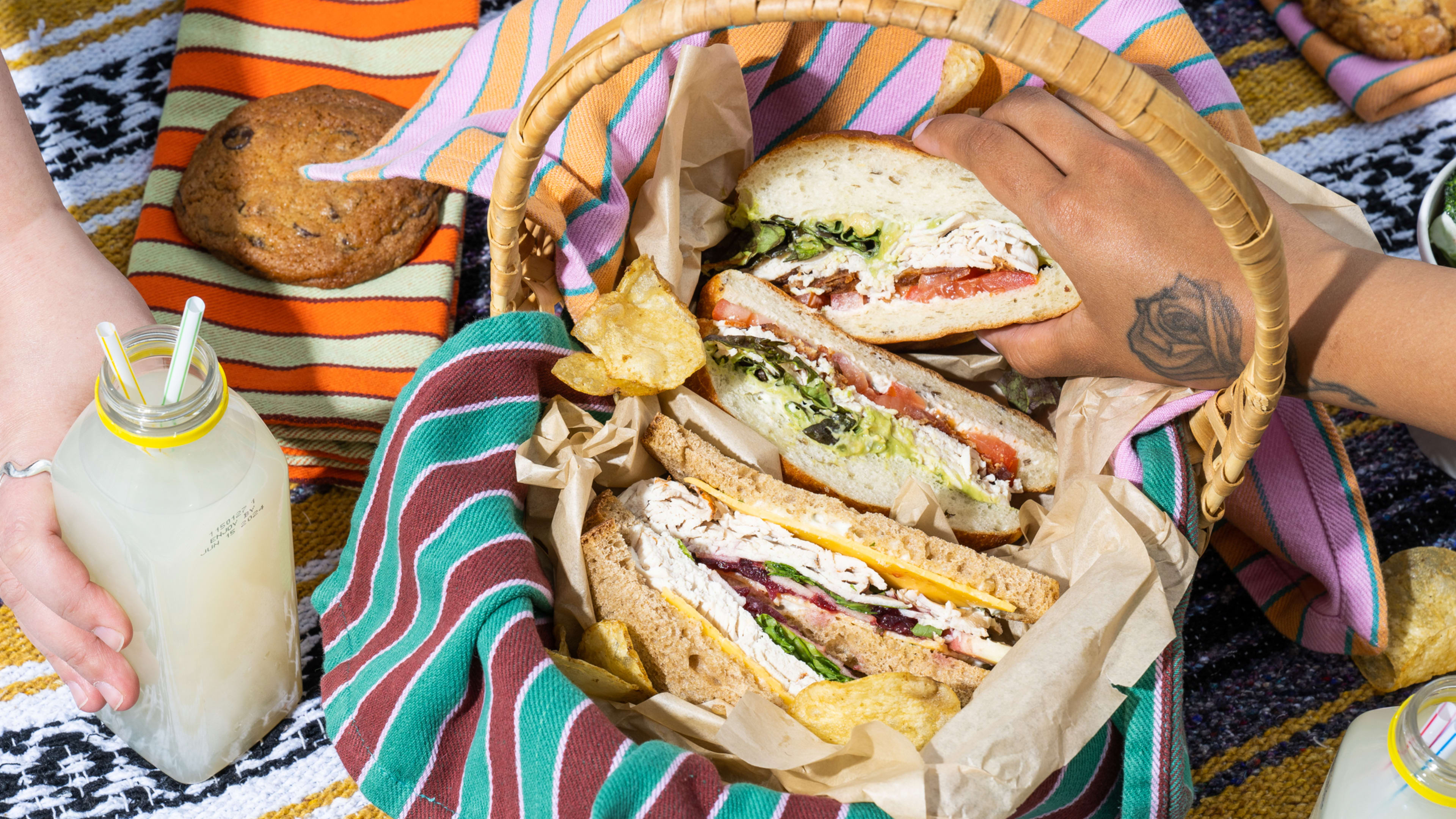 A sandwich on a picnic blanket with hands holding drinks