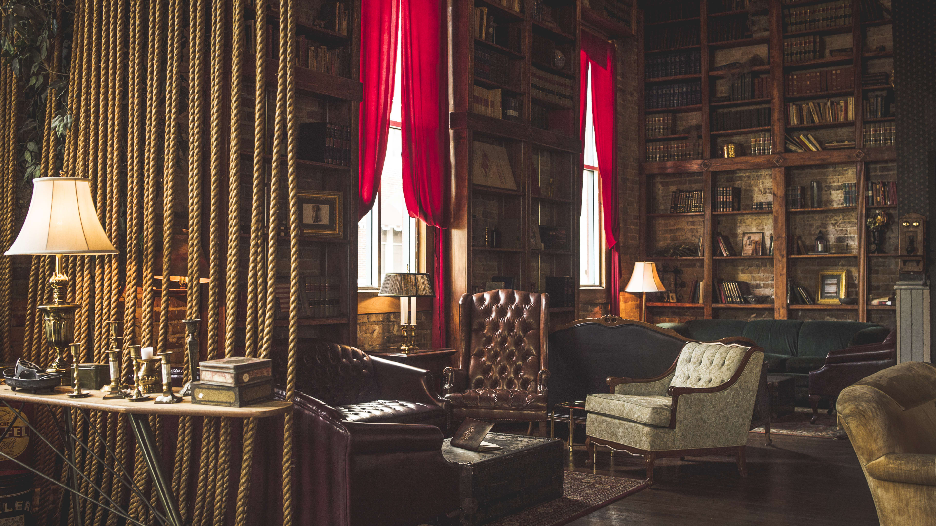 Mathers Social Gathering interiors with a large bookcase wall, mix matched cushioned chairs and couches, and large windows with long red curtains