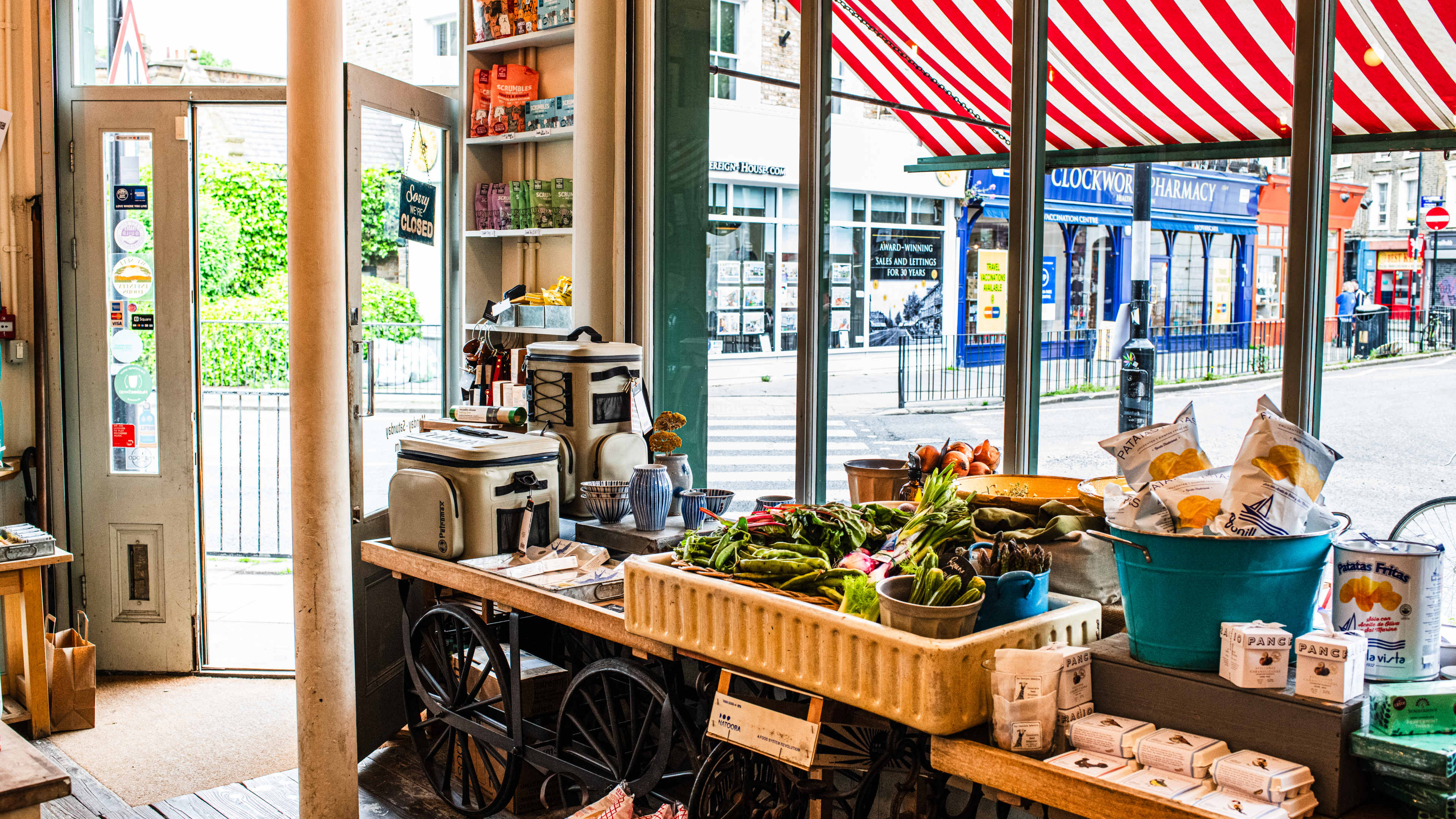 The interior of a deli with wooden benches filled with fresh vegetables and packets of crisps, with a red and white striped awning visible through the window.