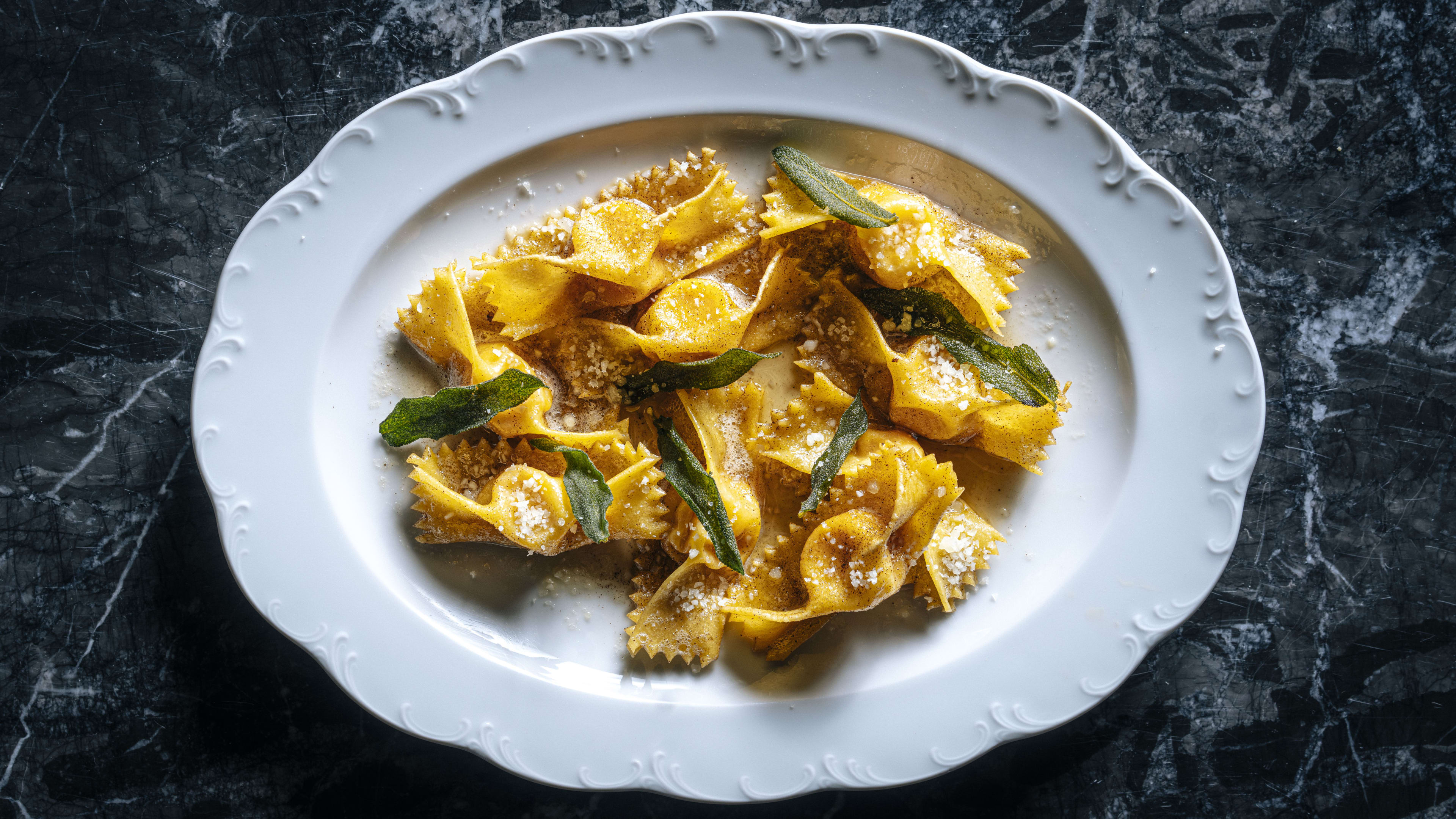 Tortelli topped with sage leaves on a scalloped white plate against a black marble background