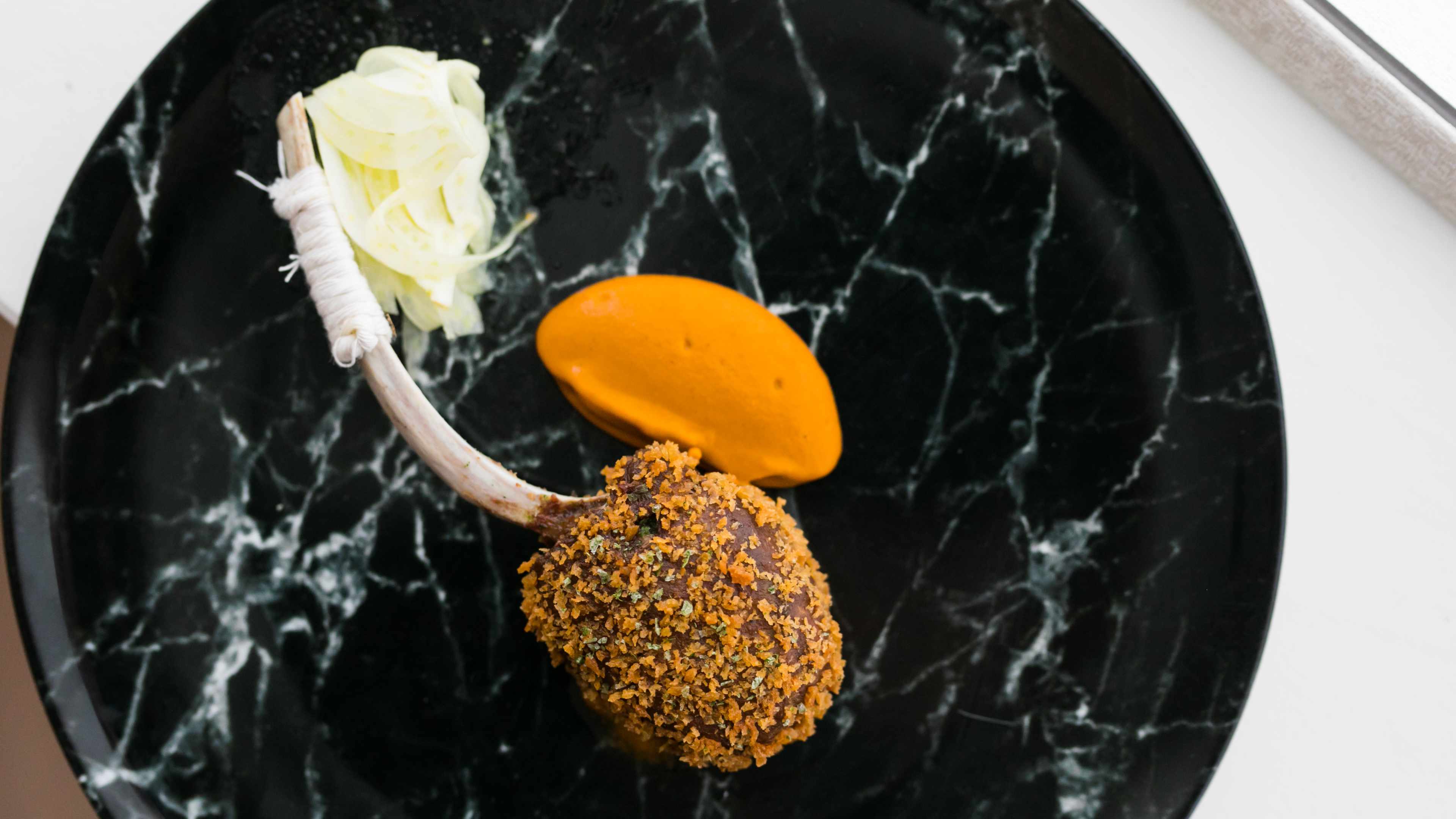 Black marble plate with artfully plated food that looks like orange sorbet and a lamb chop