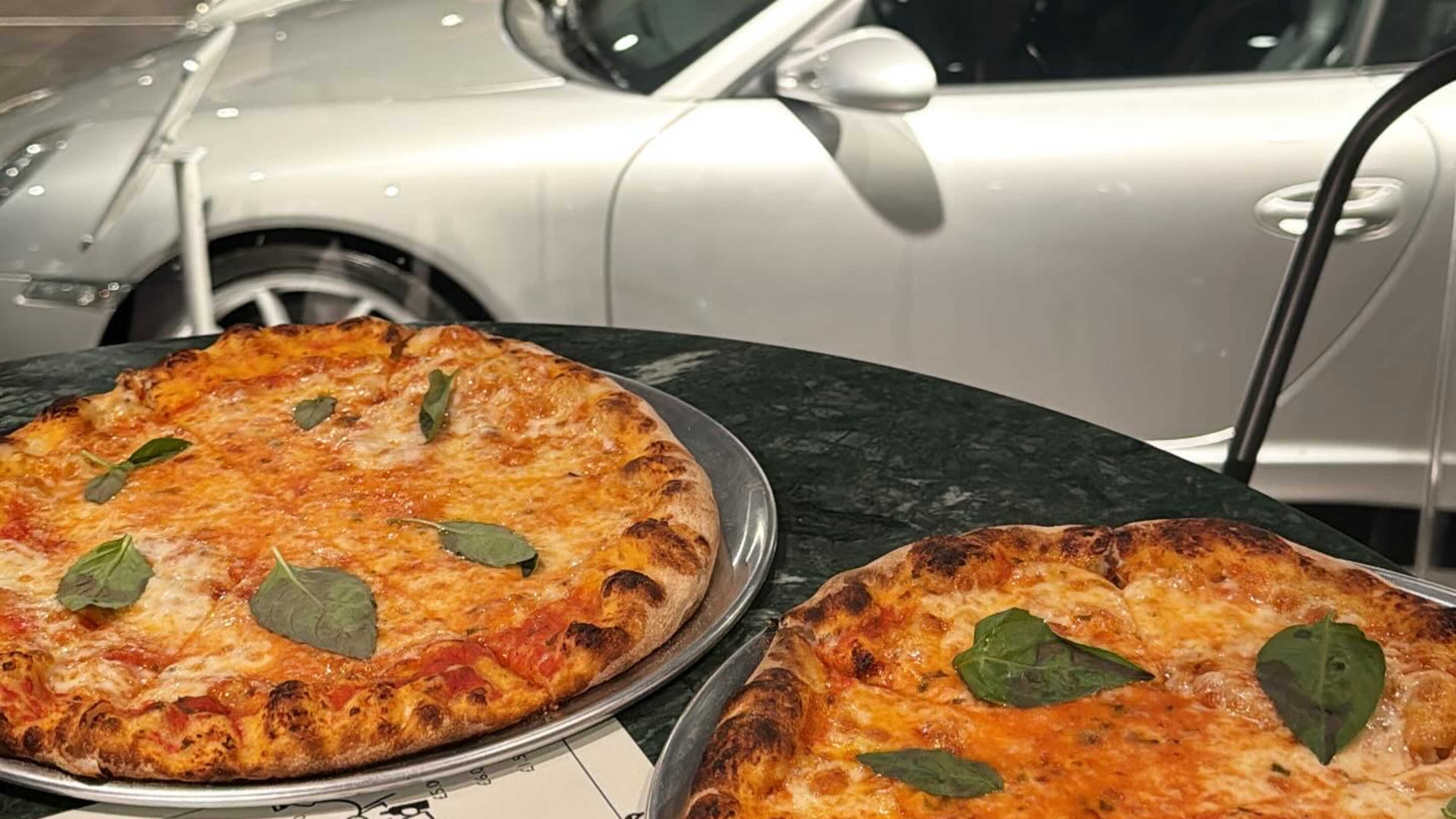 Two pizzas on a table, with vintage cars in the background.