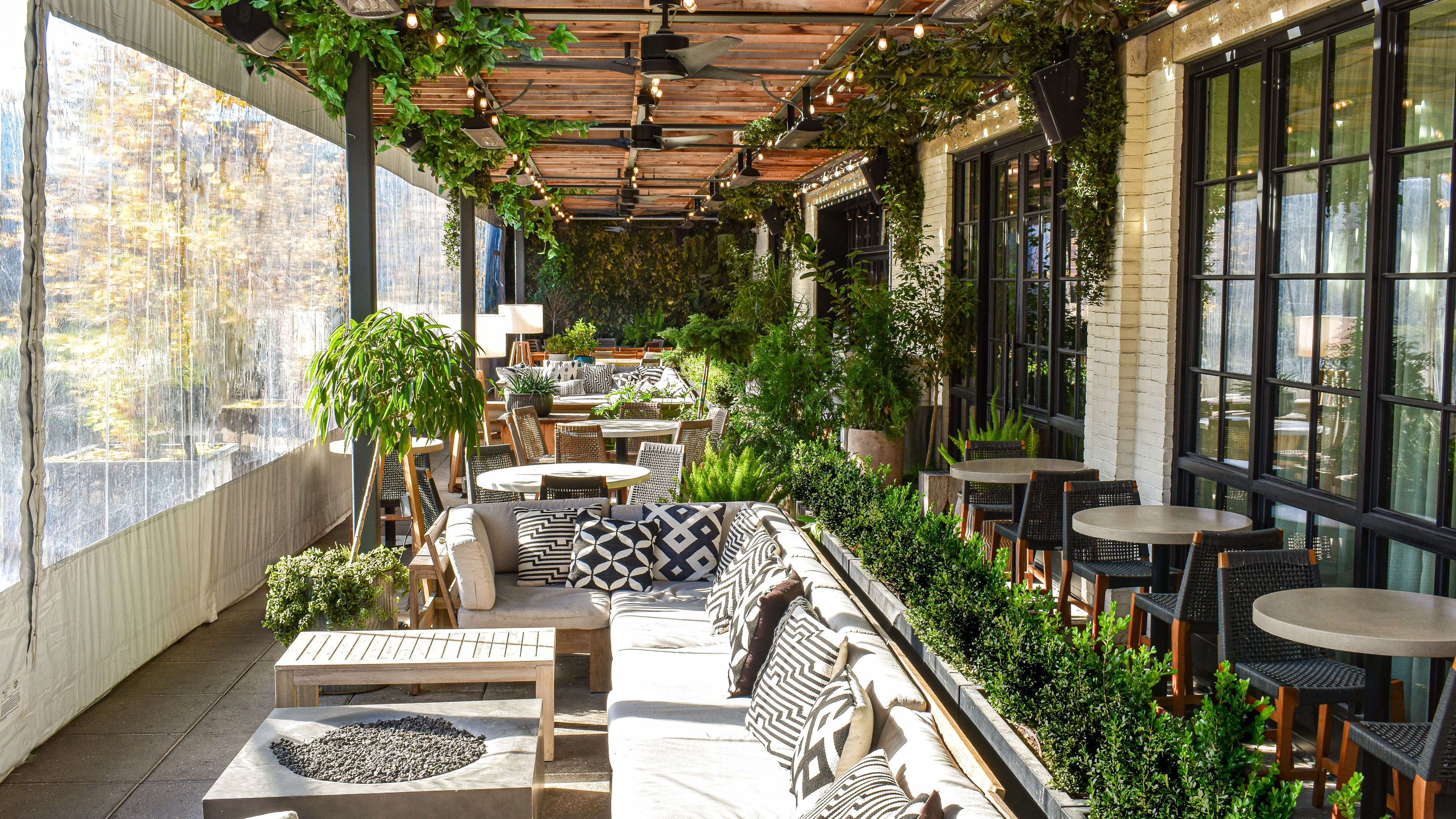 The enclosed patio at Aba. There are long couches with geometrically patterned pillows, plants lining the backs of the couches, and small tables with black woven chairs.