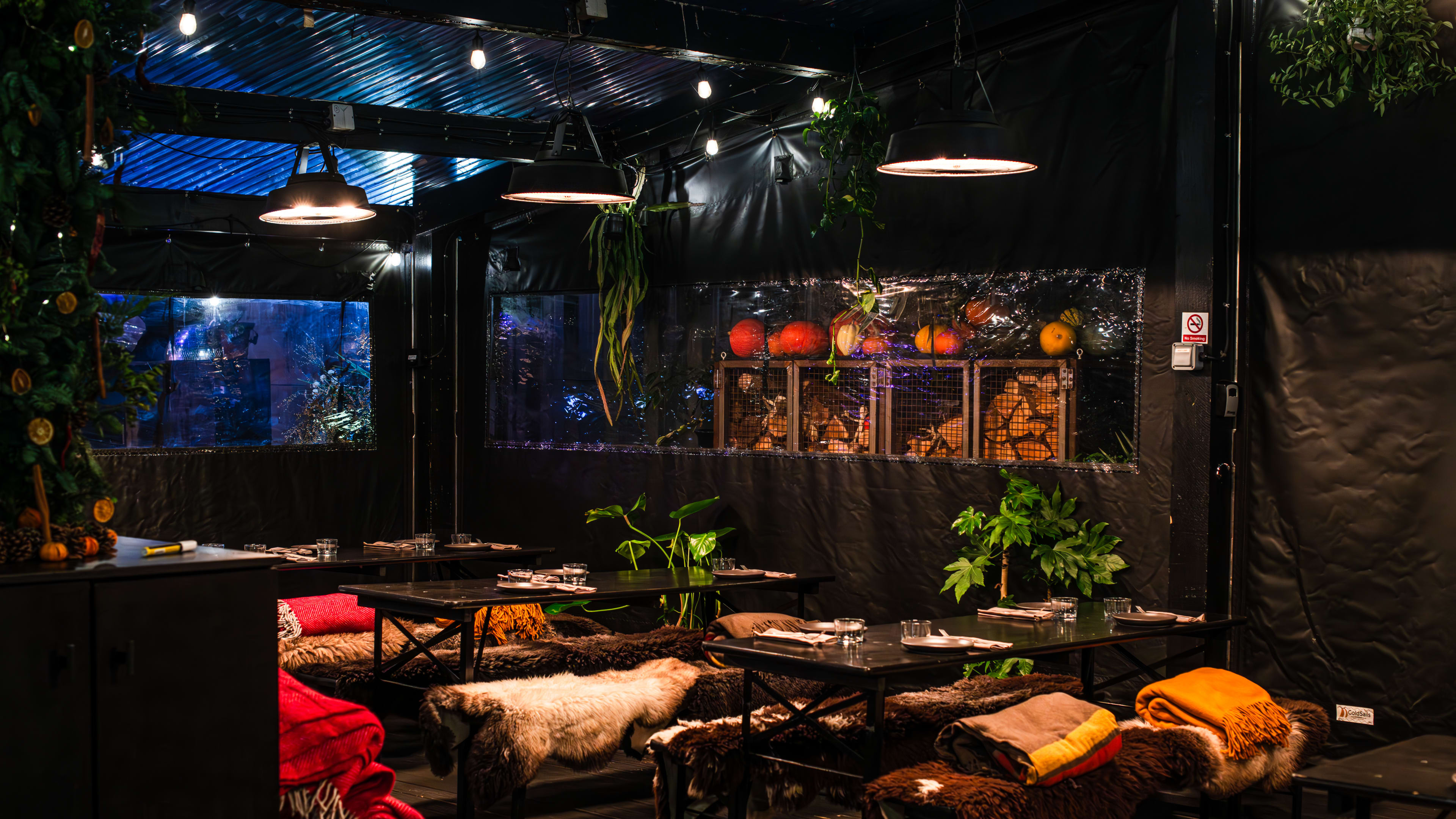 The heated, enclosed terrace at Acme Fire Cult. There are black tables and and blankets are draped over the chairs.  There are storage cubes seen in the back with logs of wood inside.