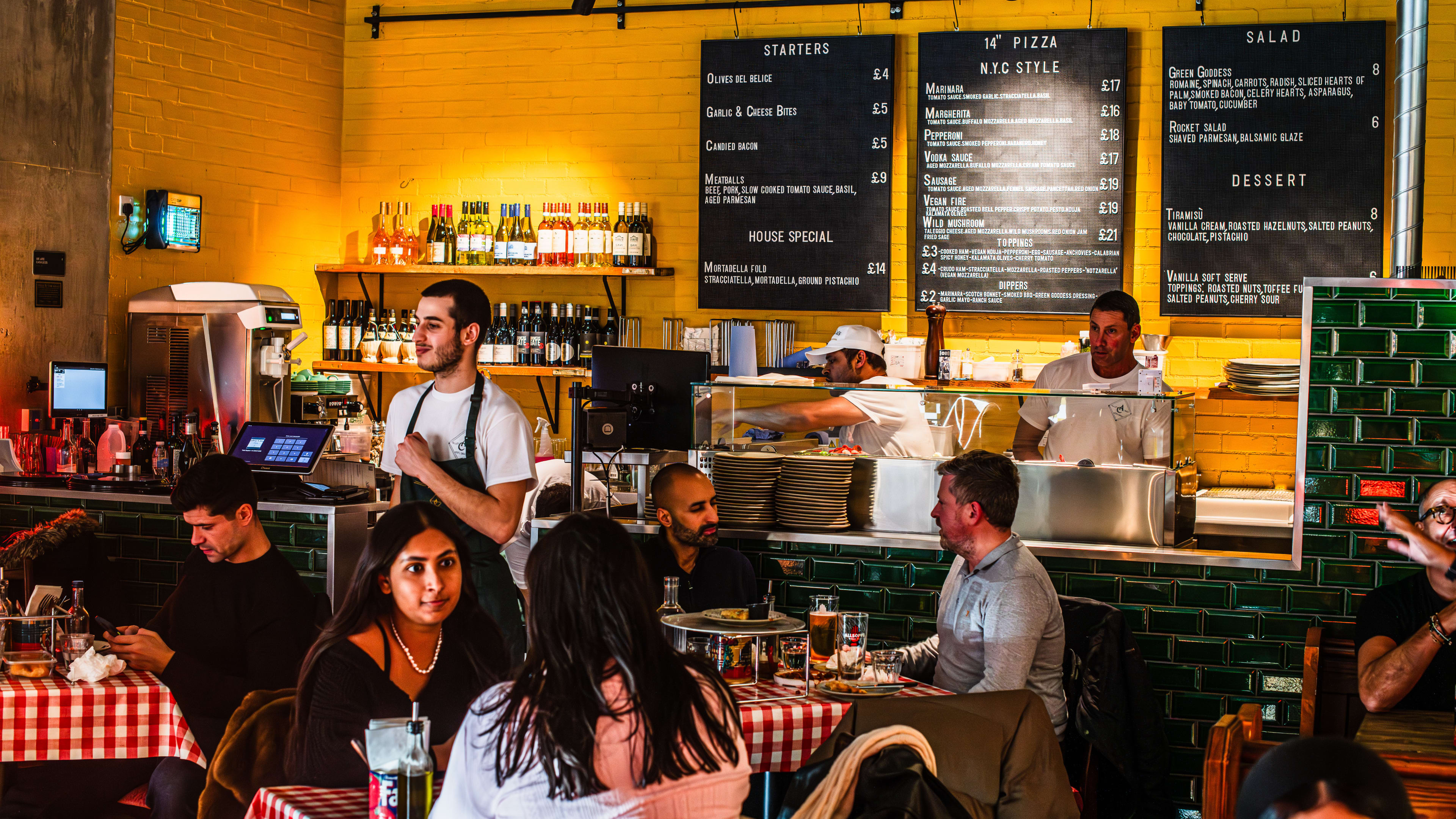 The interior of Alley Cats Pizza with people sitting at square tables with checkered tablecloths. Menu items and specials are listed on boards on the wall behind the kitchen counter.