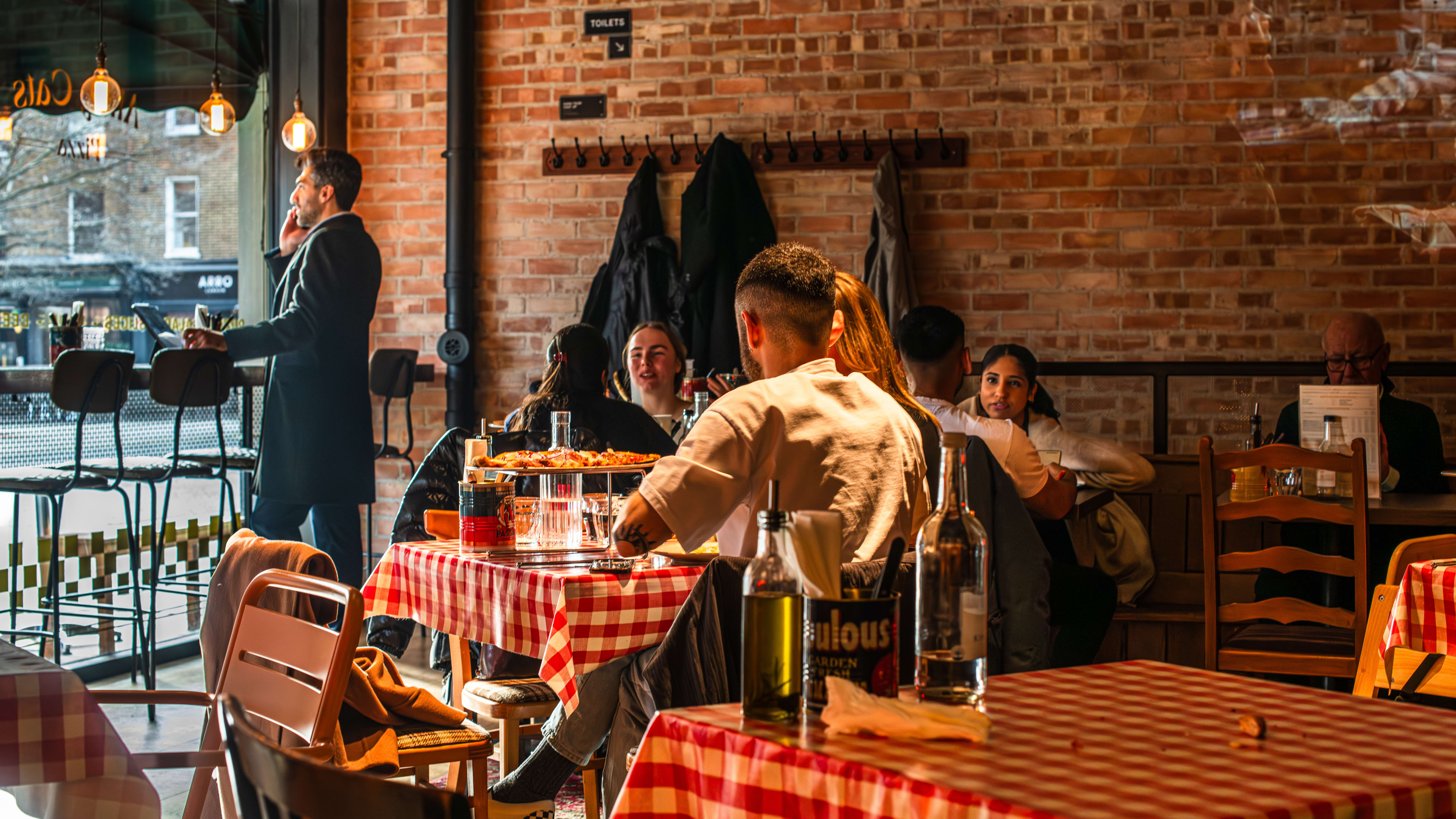 The brick walled interior of Alley Cats Pizza with people sitting at square tables with checkered tablecloths and a person standing at the window counter on the phone.