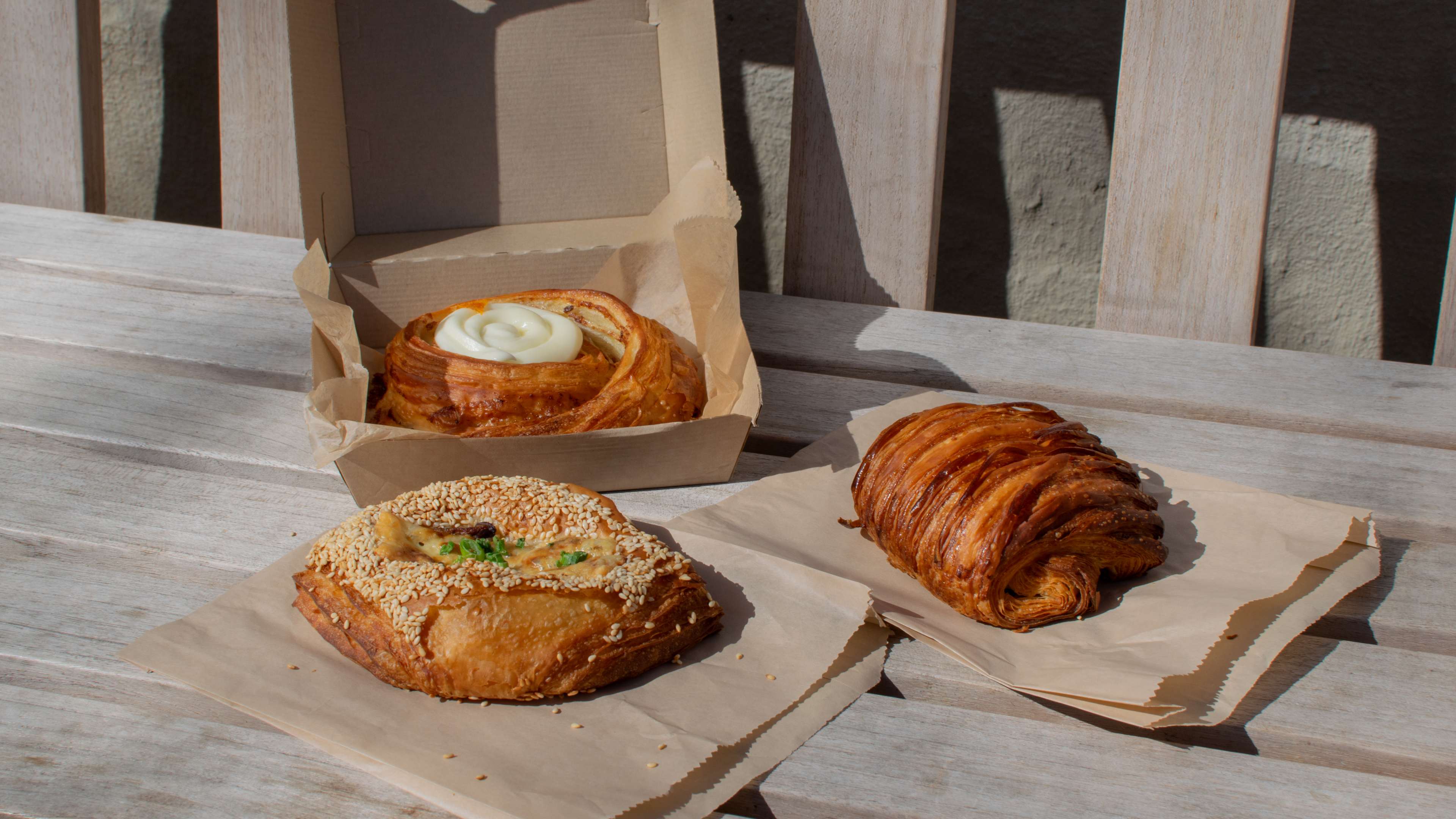 A cinnamon roll, savory pastry, and chocolate croissant on a park bench.