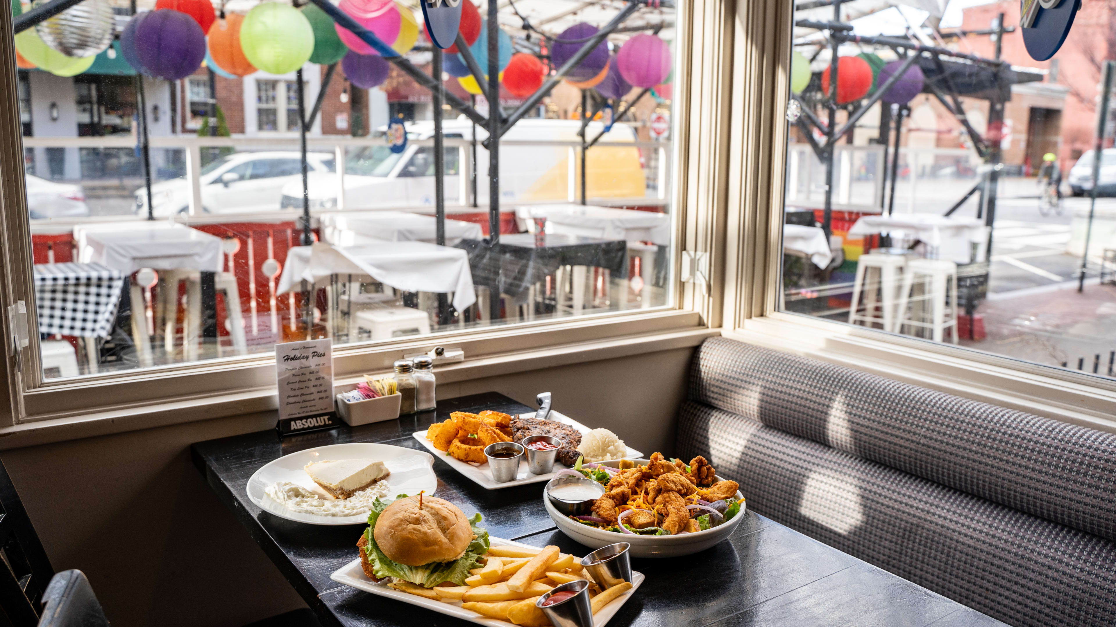 Burger, cheesecake, and salad on table near window overlooking patio where rainbow lanterns are hung.