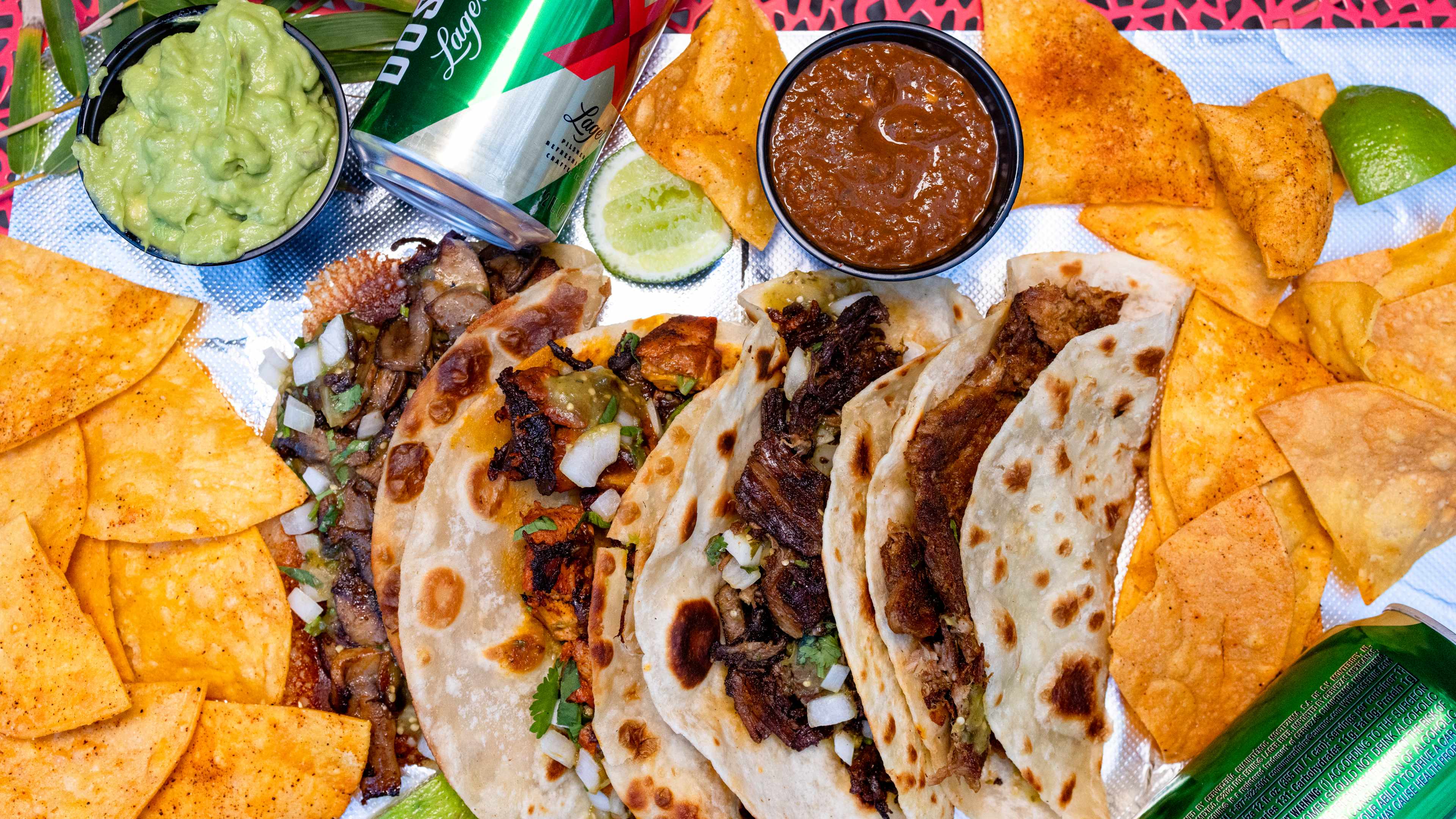 Four different tacos on foil surrounded with tortilla chips, sliced limes, a small bowl of guacamole and two cans of Dos Equis.