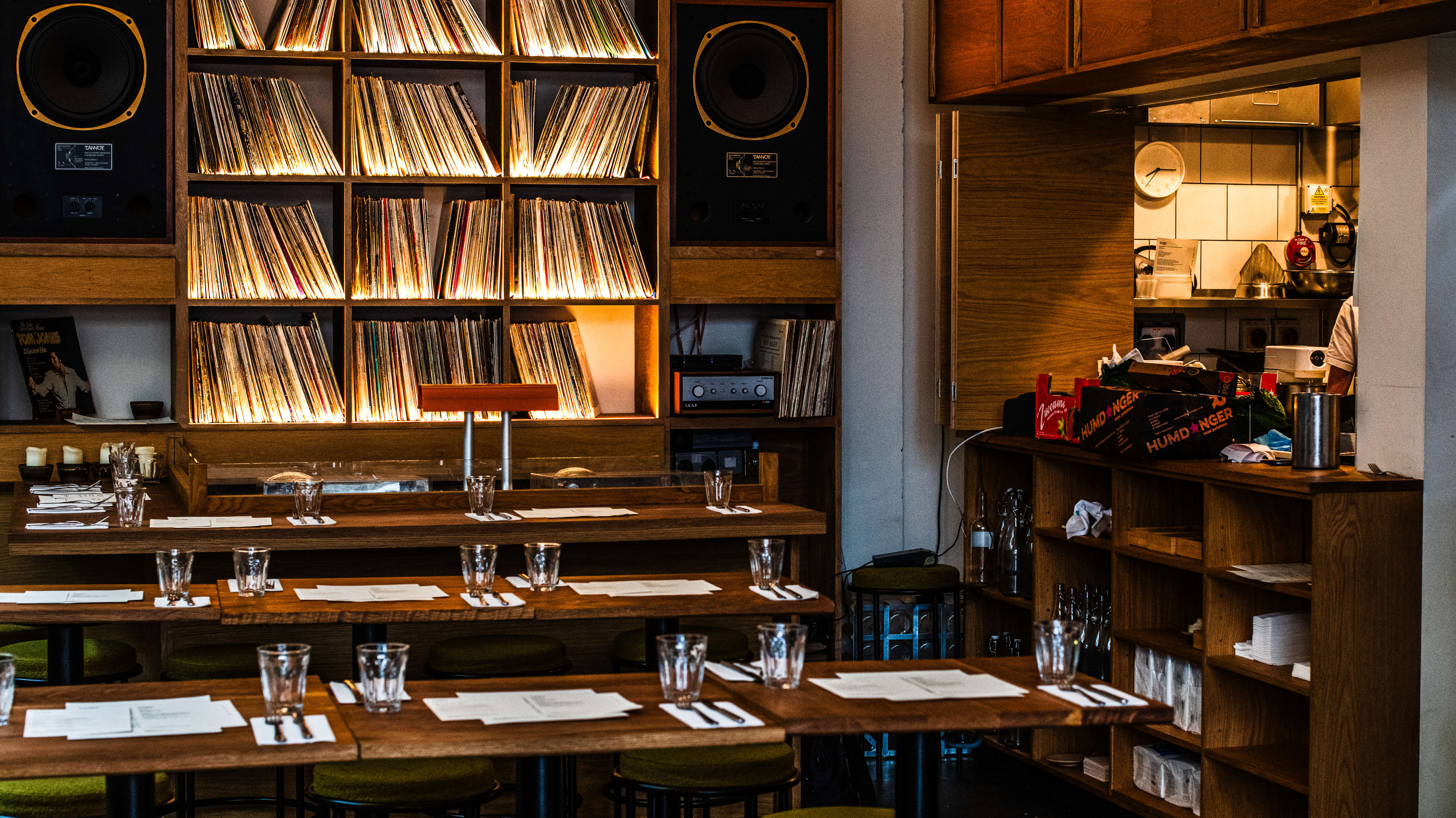 The interior of Bambi with long wooden tables with green chairs, an open kitchen to the right and vinyl records lining the shelves on the back wall with two large speakers.