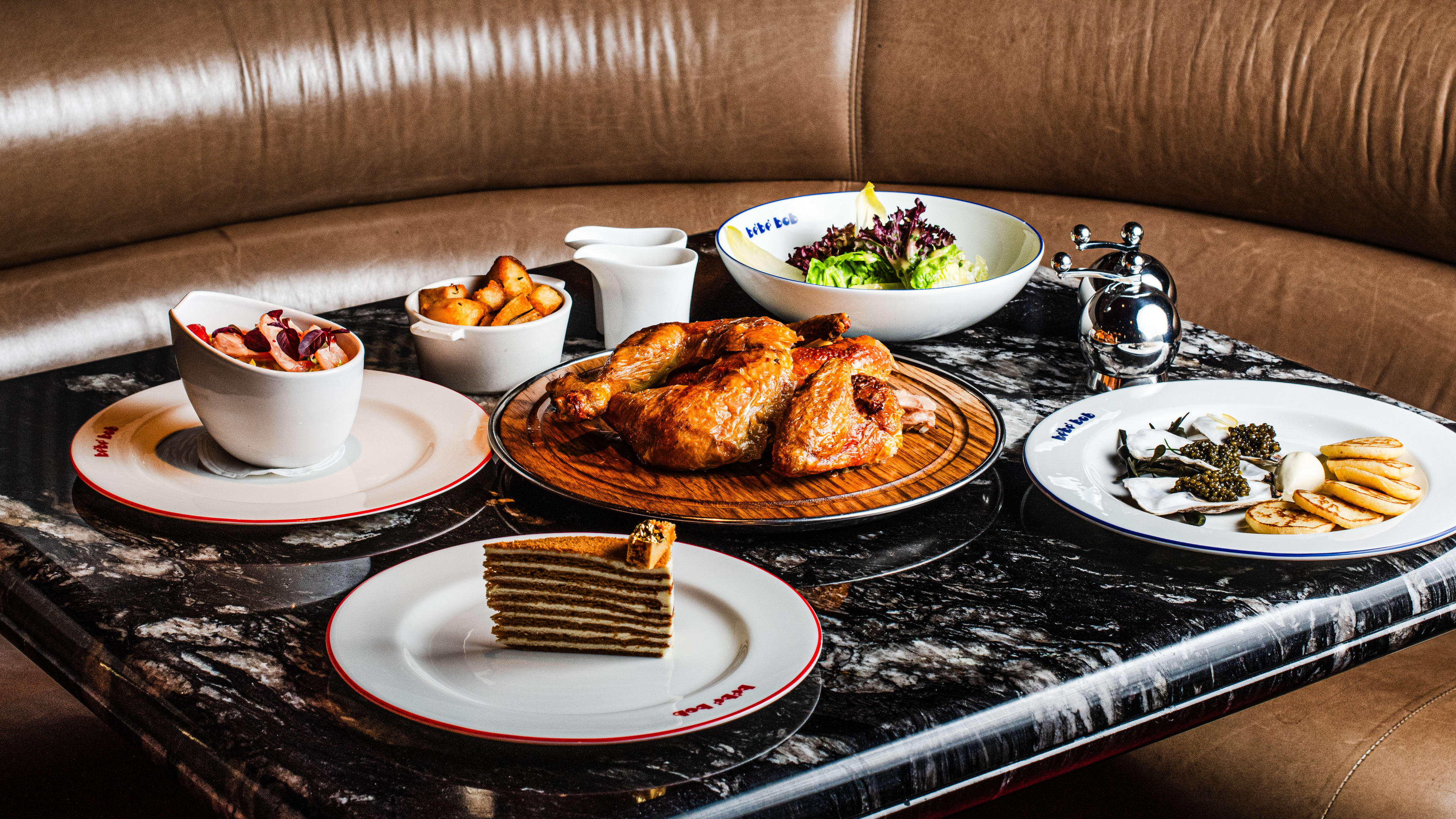 A spread of dishes on white dish ware on a black marble table surrounded by a brown leather booth.