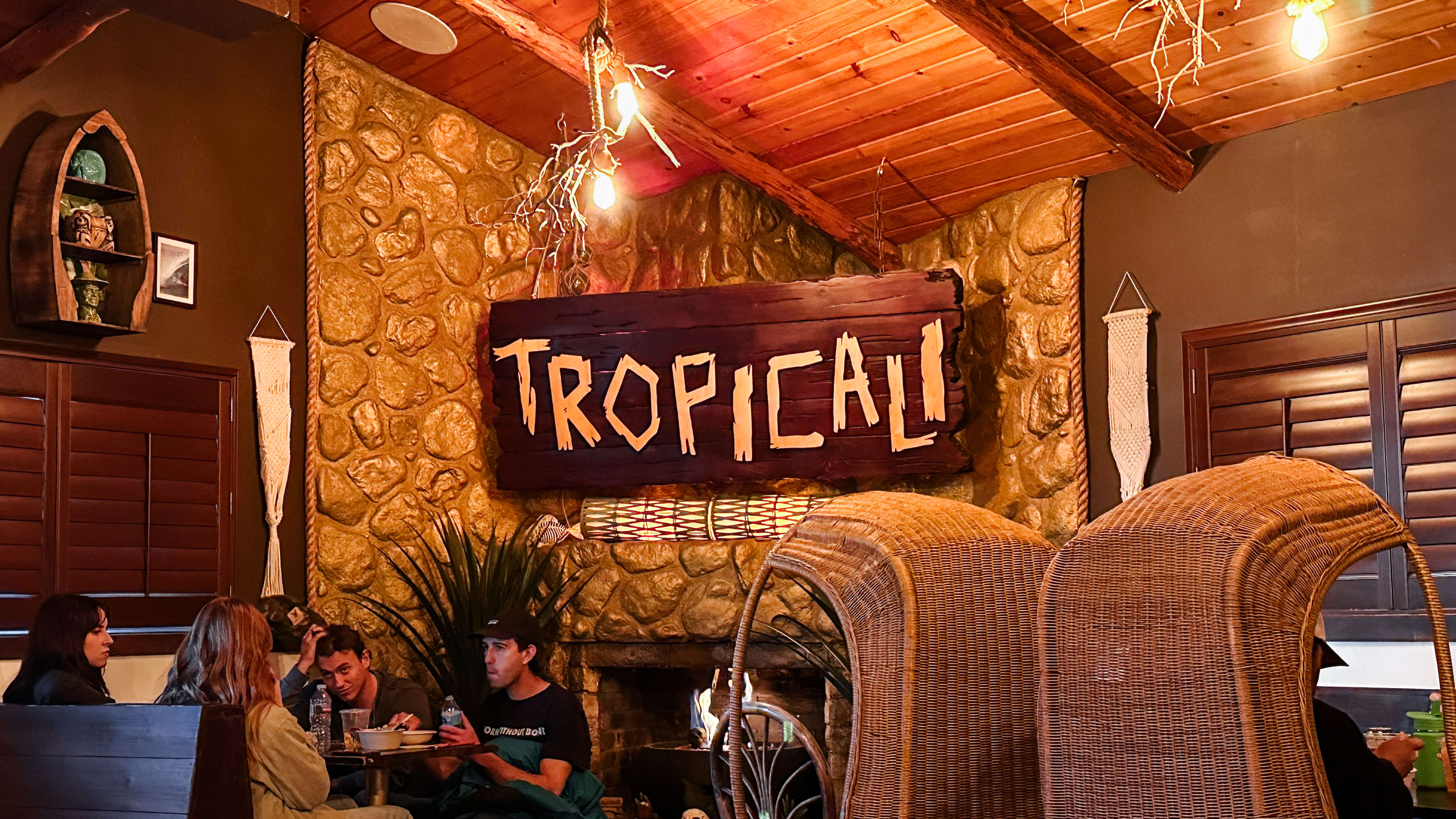 The wooden and stone interior of Tropicali. A large wooden sign, reading "Tropicali" hangs above a fireplace. There are large wicker egg chairs and wooden booths.
