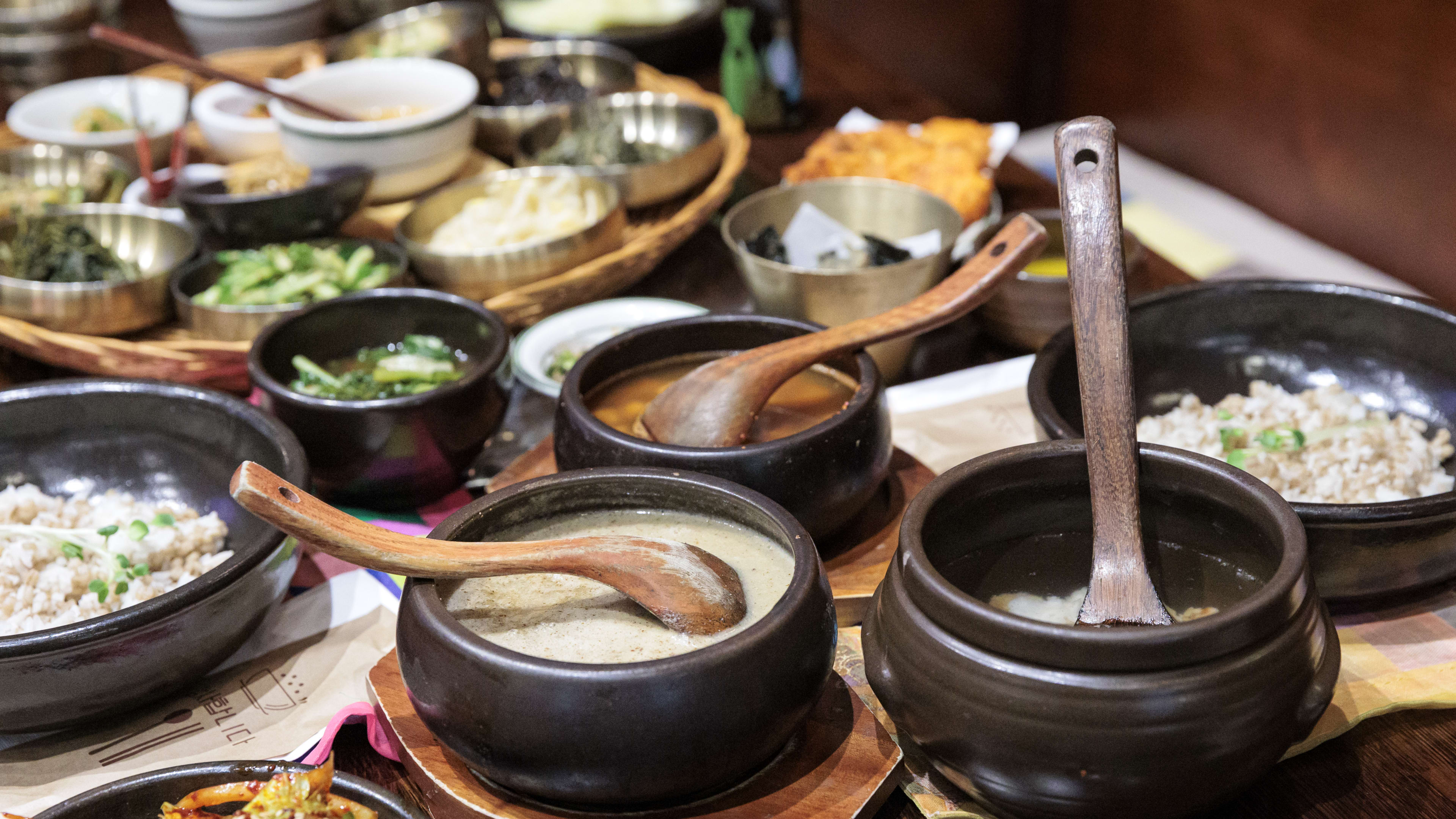 A large spread of set menu dishes and sides in ceramic and metal bowls with wooden ladles on a wooden table.