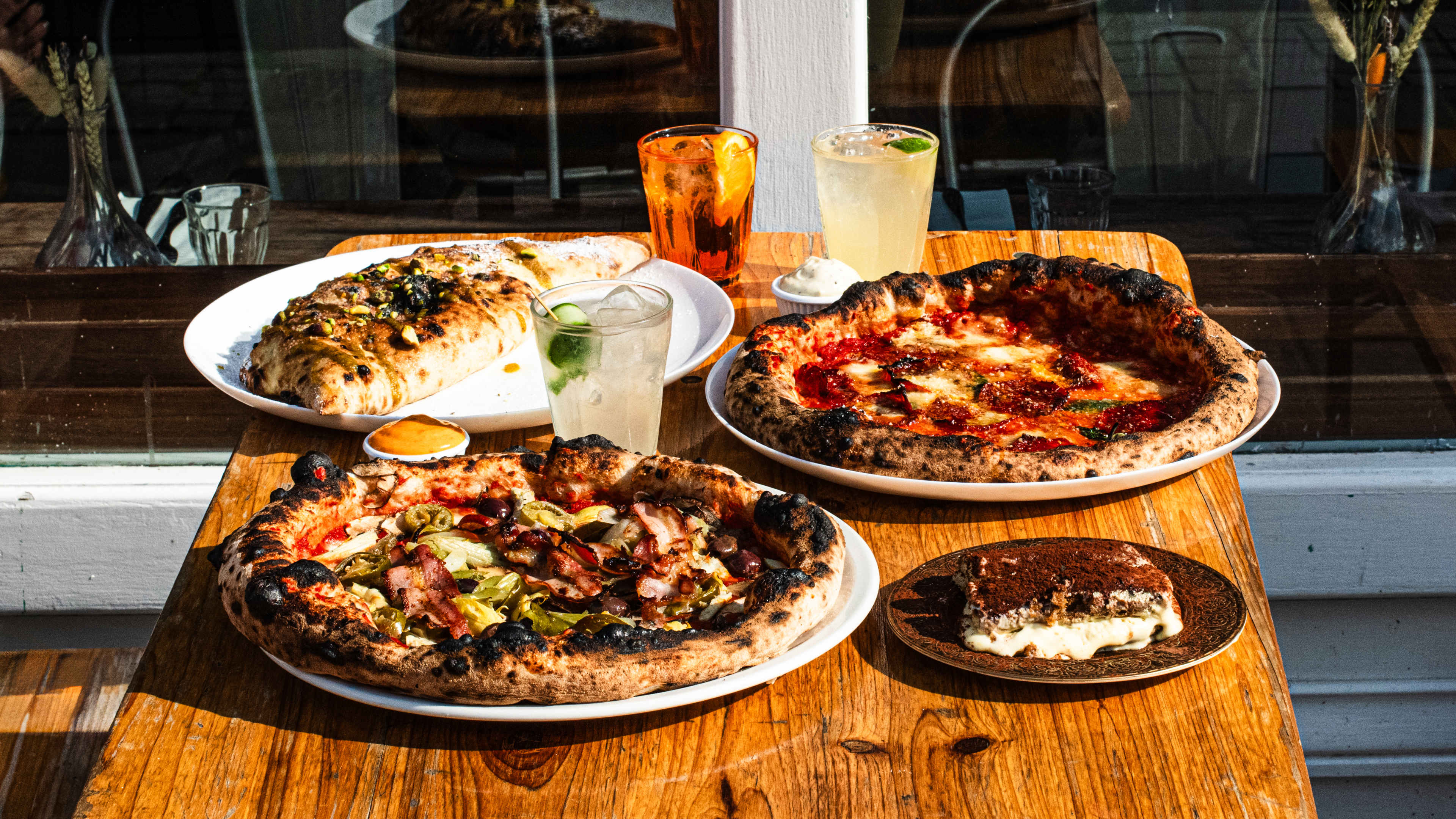 Two pizzas, a tiramisu, drinks, and a calzone on a wooden outside table in the sunshine.