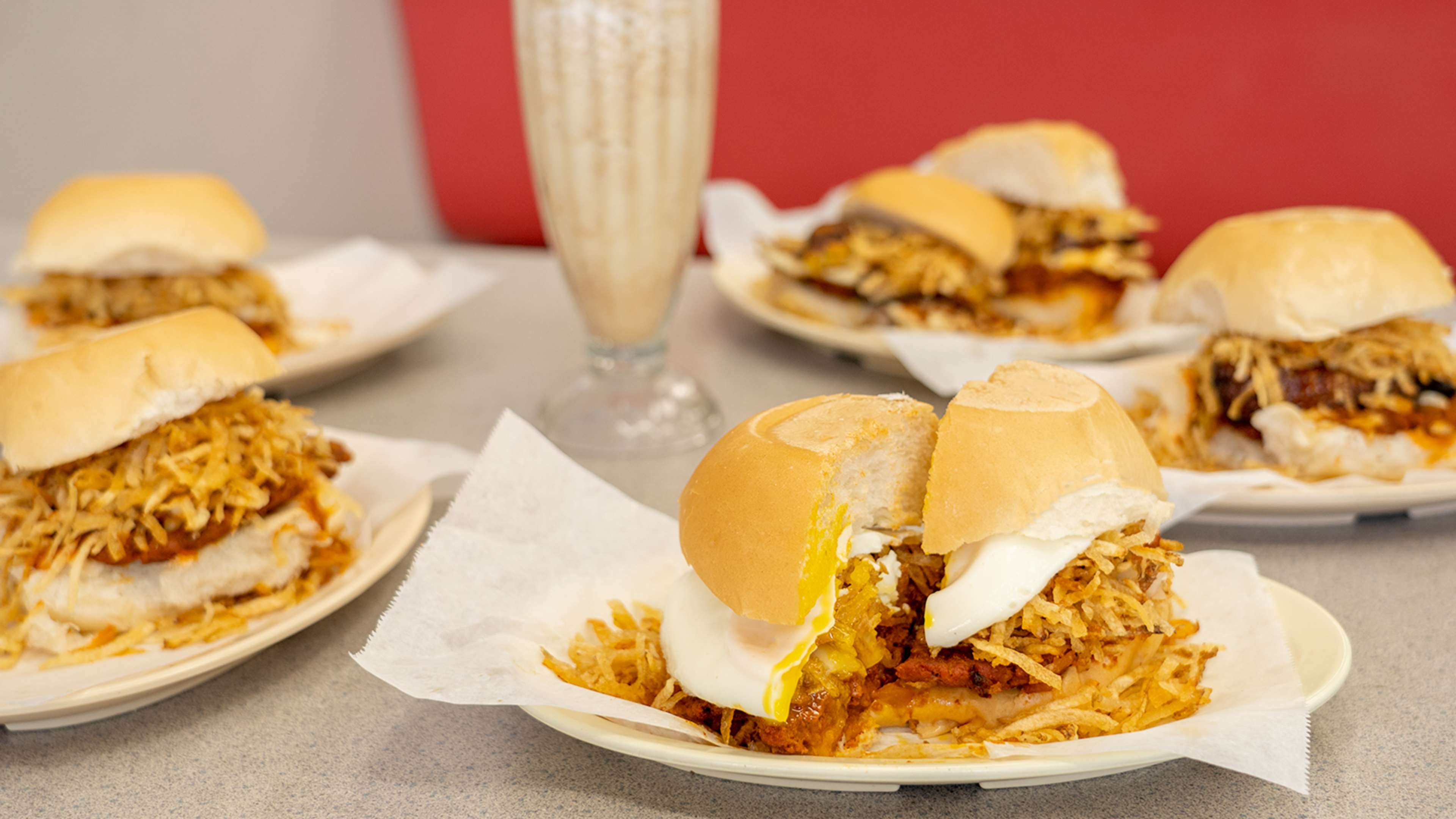 Array of sandwiches with fried potatoes and eggs next to a milkshake in a classic diner glass.