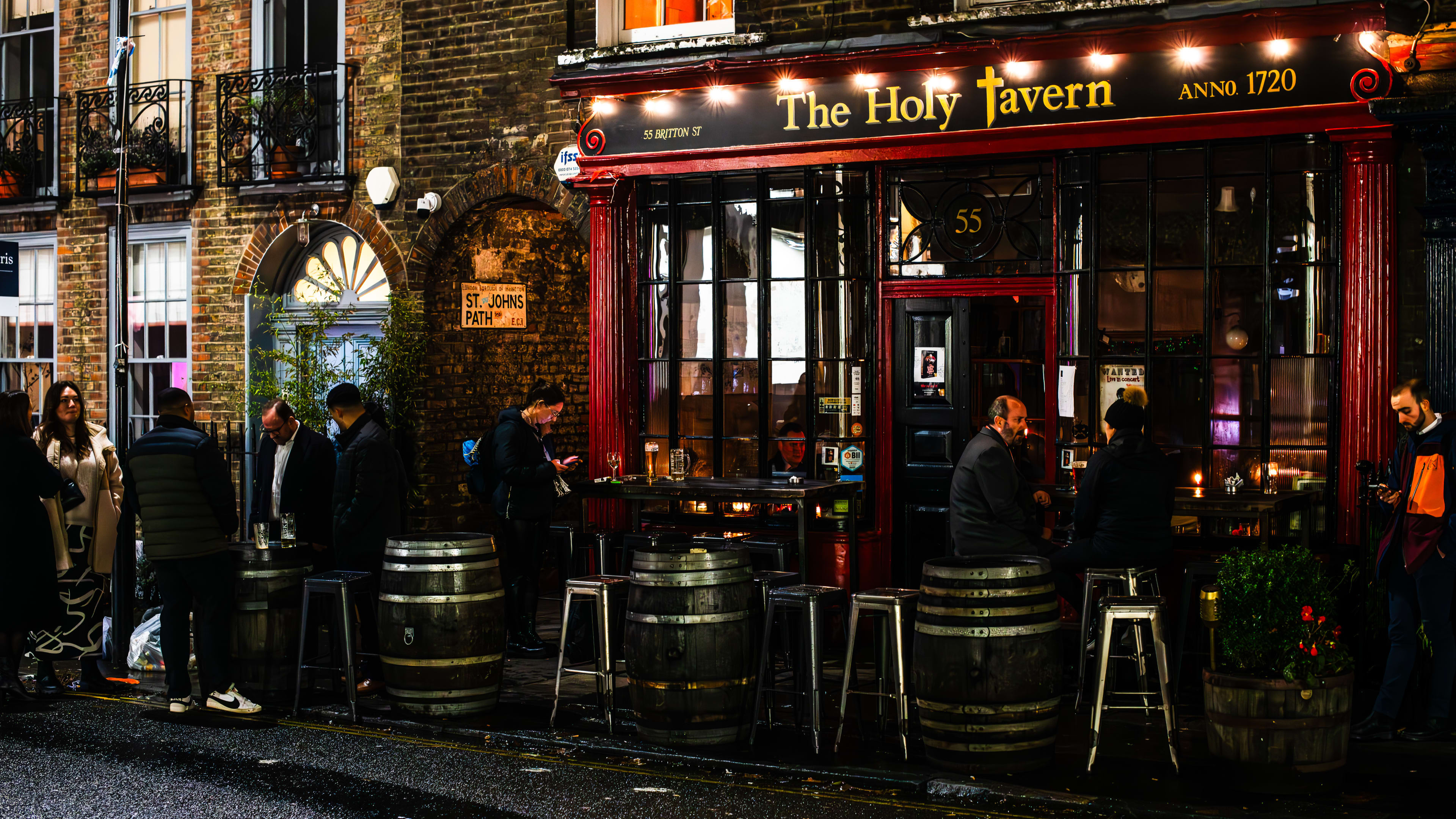 The bustling exterior of The Holy Tavern, with people sitting and standing around barrel tables and metal barstools. The inside of the bar is only lit by candles.