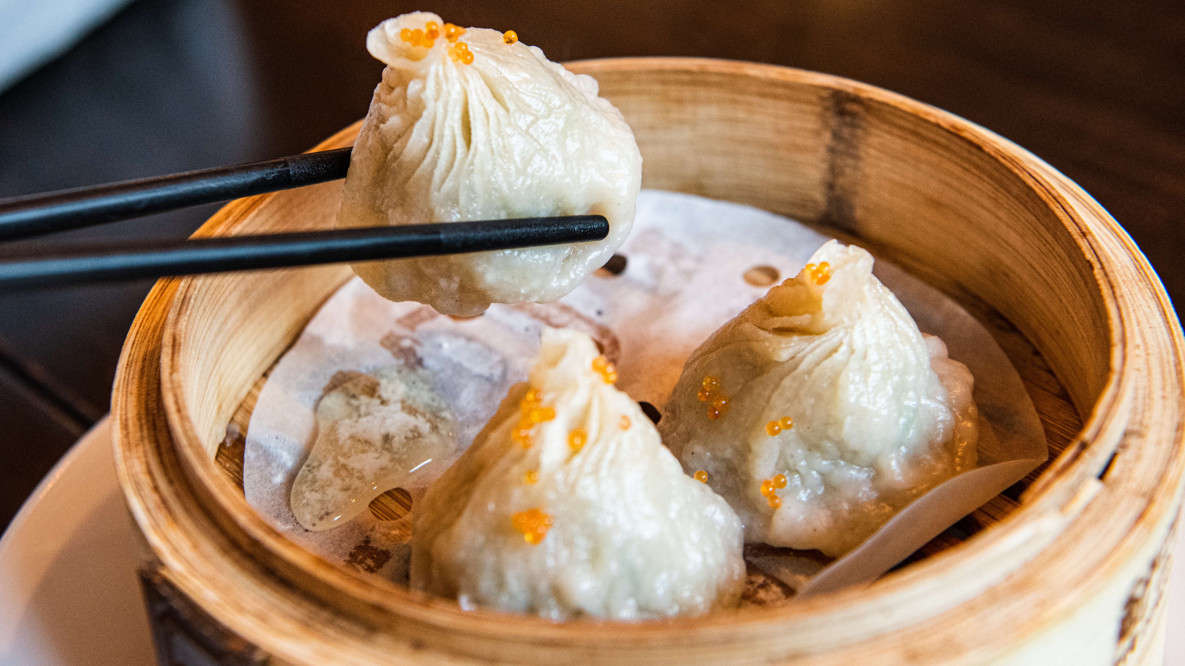 black chopsticks holding up a crab xiao long bao from a steamer basket filled with two more. They are all sprinkled with tobiko.