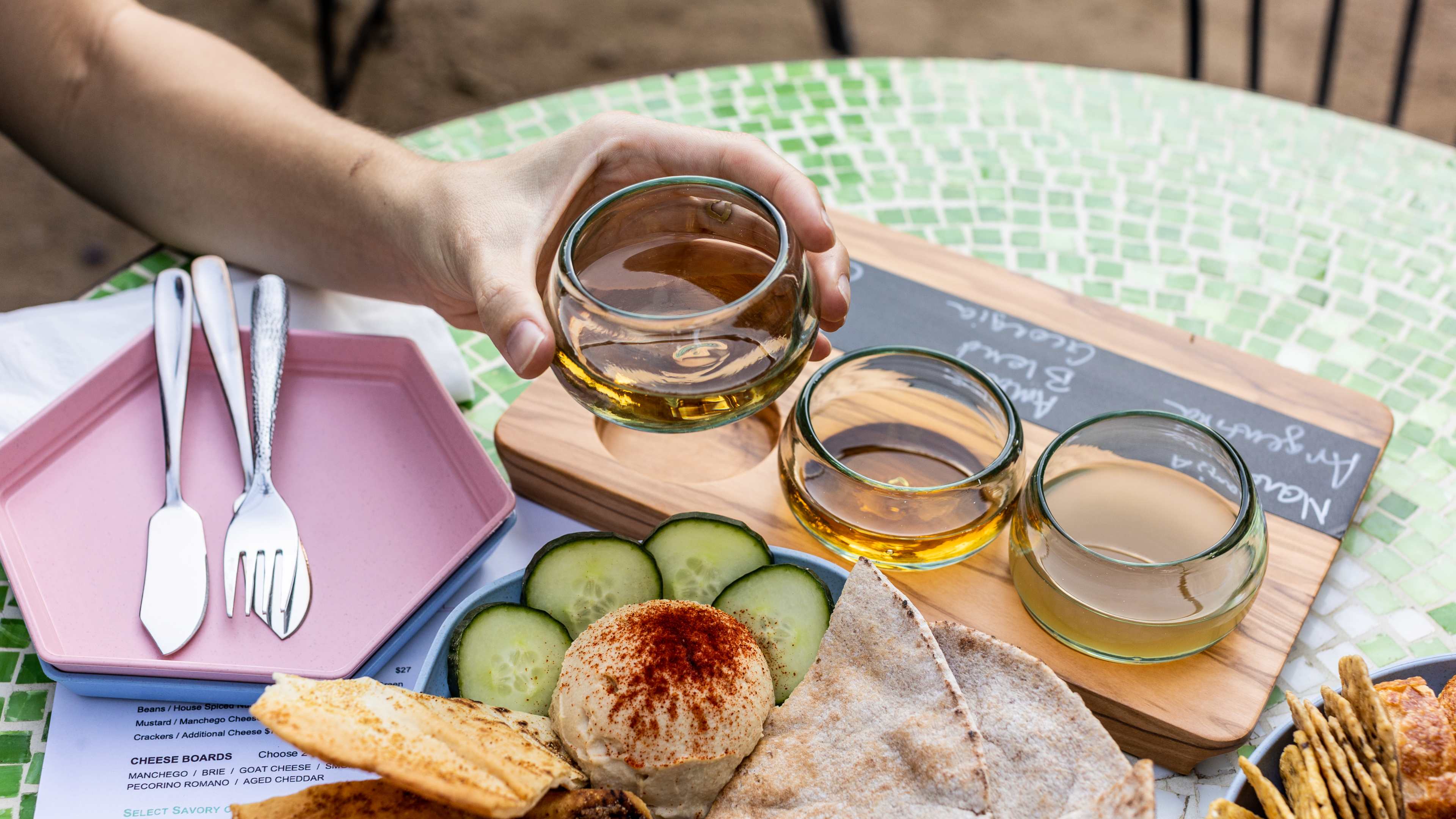 A person holding up a glass from the Orange Wine Flight wooden holder. There is a plate of pita and hummus and cucumber, with a pink hexigon shaped plate on the side. All of this is sitting on a green mosaic tiled table.