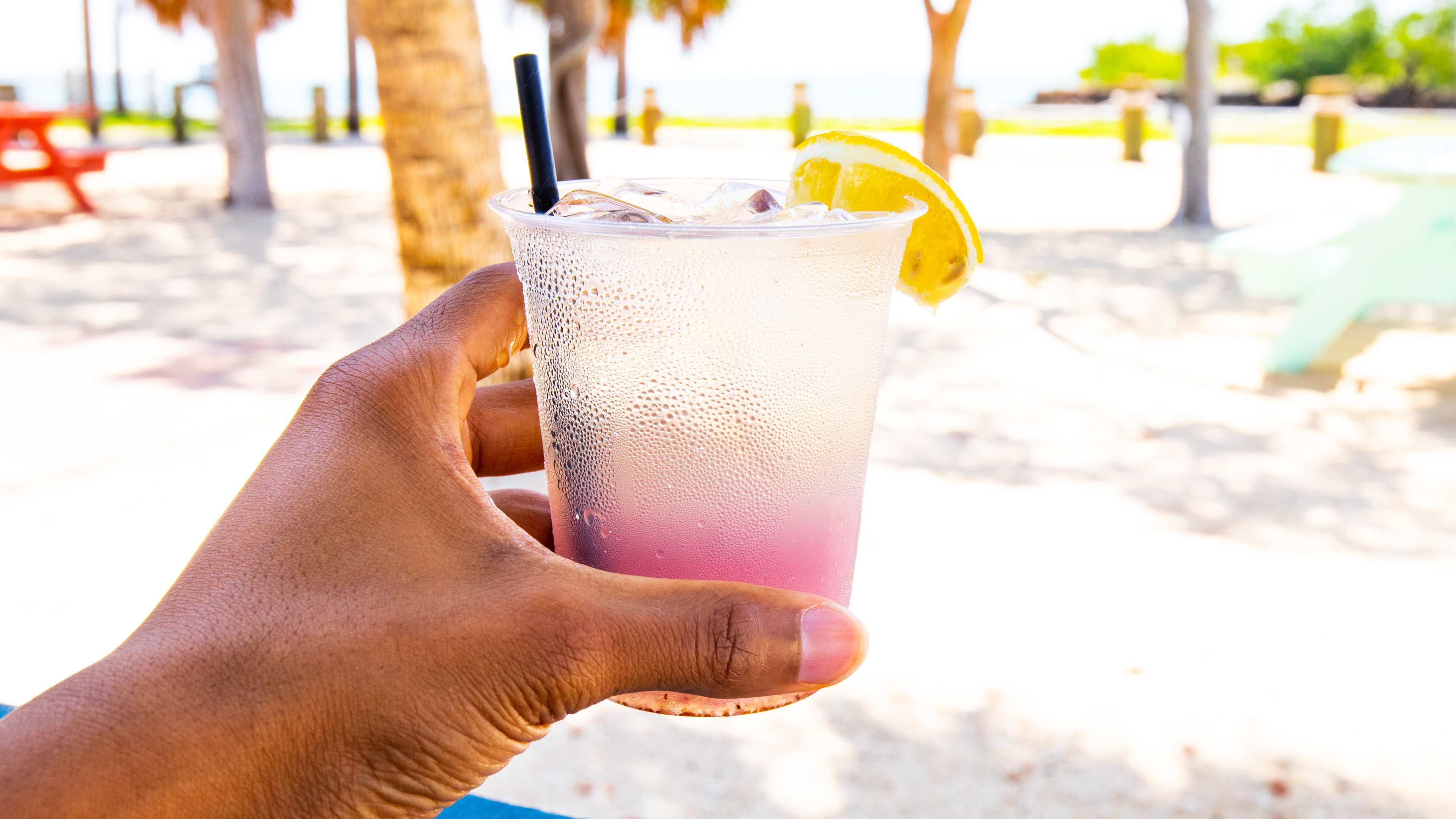 Hand holding plastic cup filled with ombre colorful drink garnished with lemon against beach background.