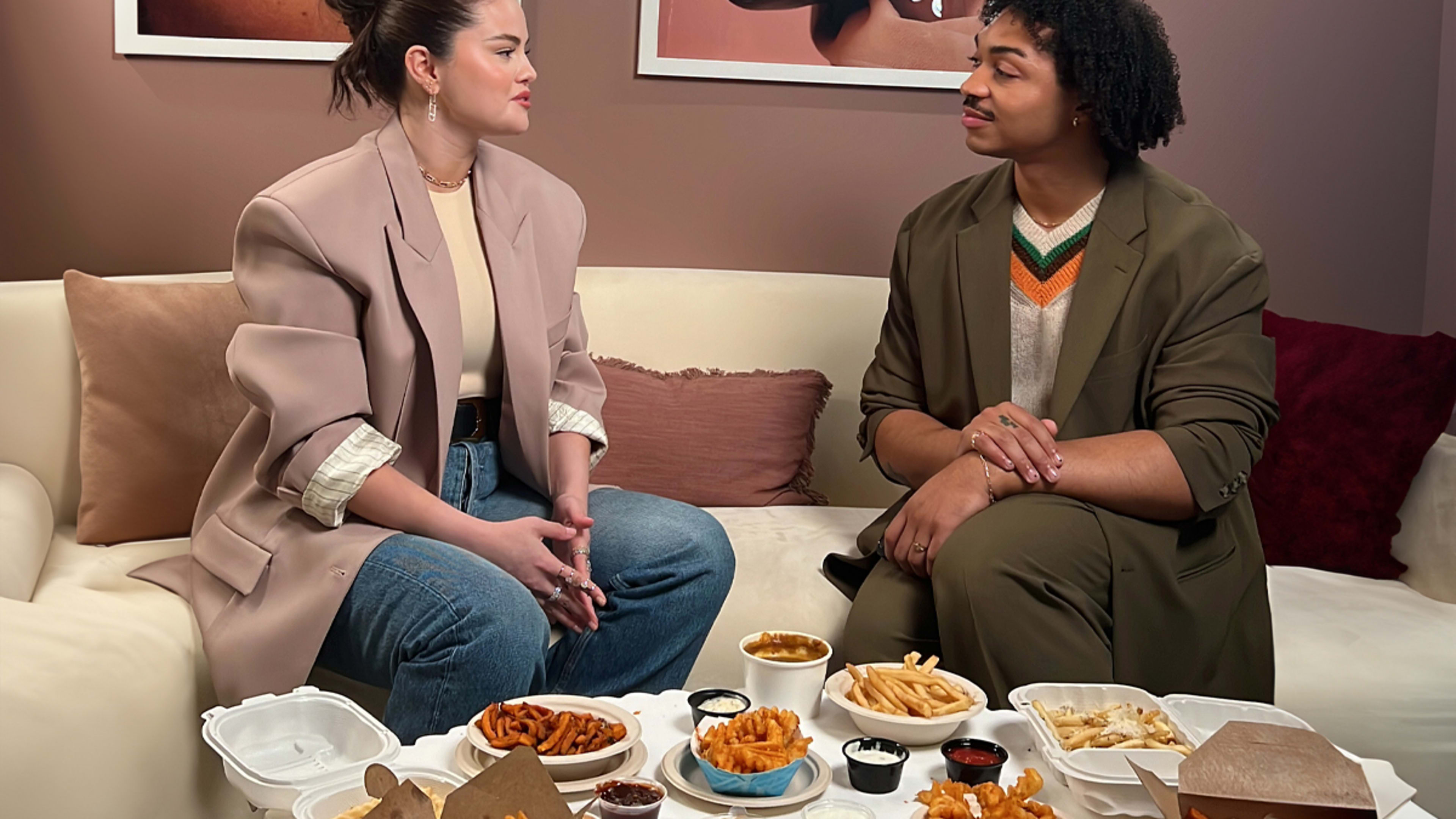 The singer Selena Gomez sits next to Infatuation writer Nikko Duren on a couch in front of a table full of French fries